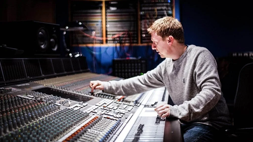 A man in a gray sweatshirt using a large audio mixing console in a music studio.