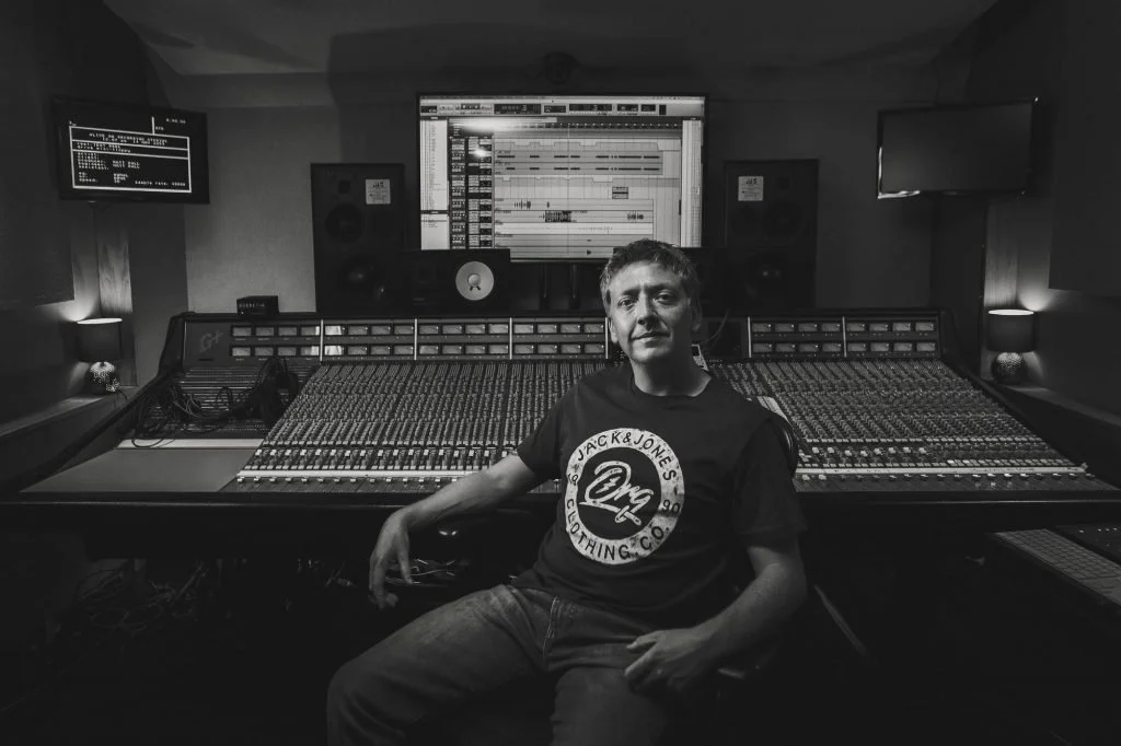 A man sitting in a recording studio with a large mixing console and digital audio workstation on a screen behind him.