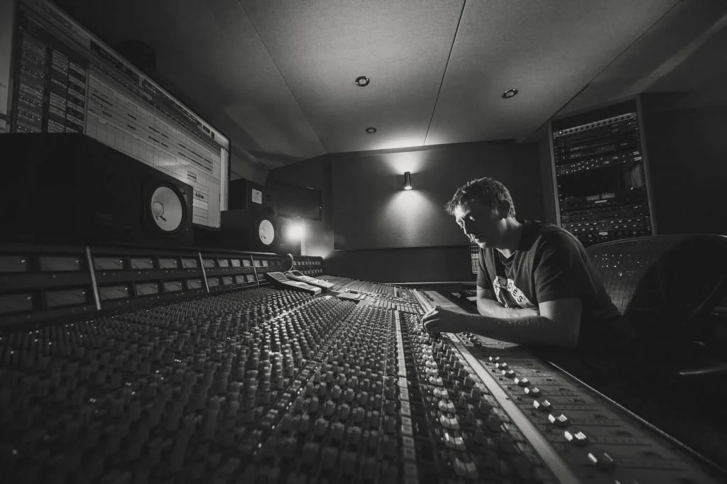 A man working at a large music mixing console in a recording studio, illuminated by soft lighting.