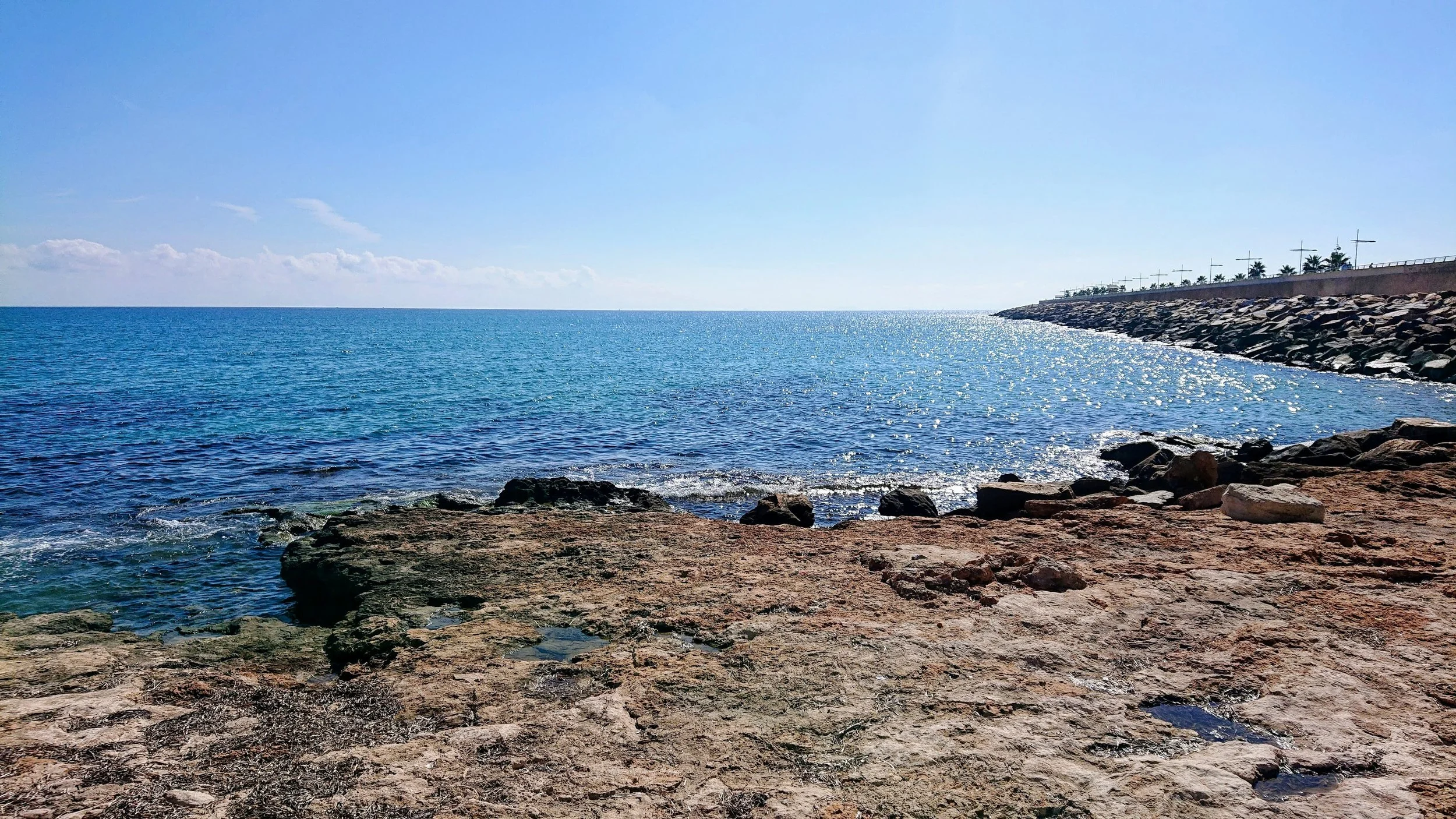 This vibrant coastal image showcases a sunlit stretch of rocky shoreline overlooking the sparkling blue waters of the Mediterranean Sea. Gentle waves shimmer under the bright sky, creating a tranquil and inviting seaside atmosphere. To the right, a l