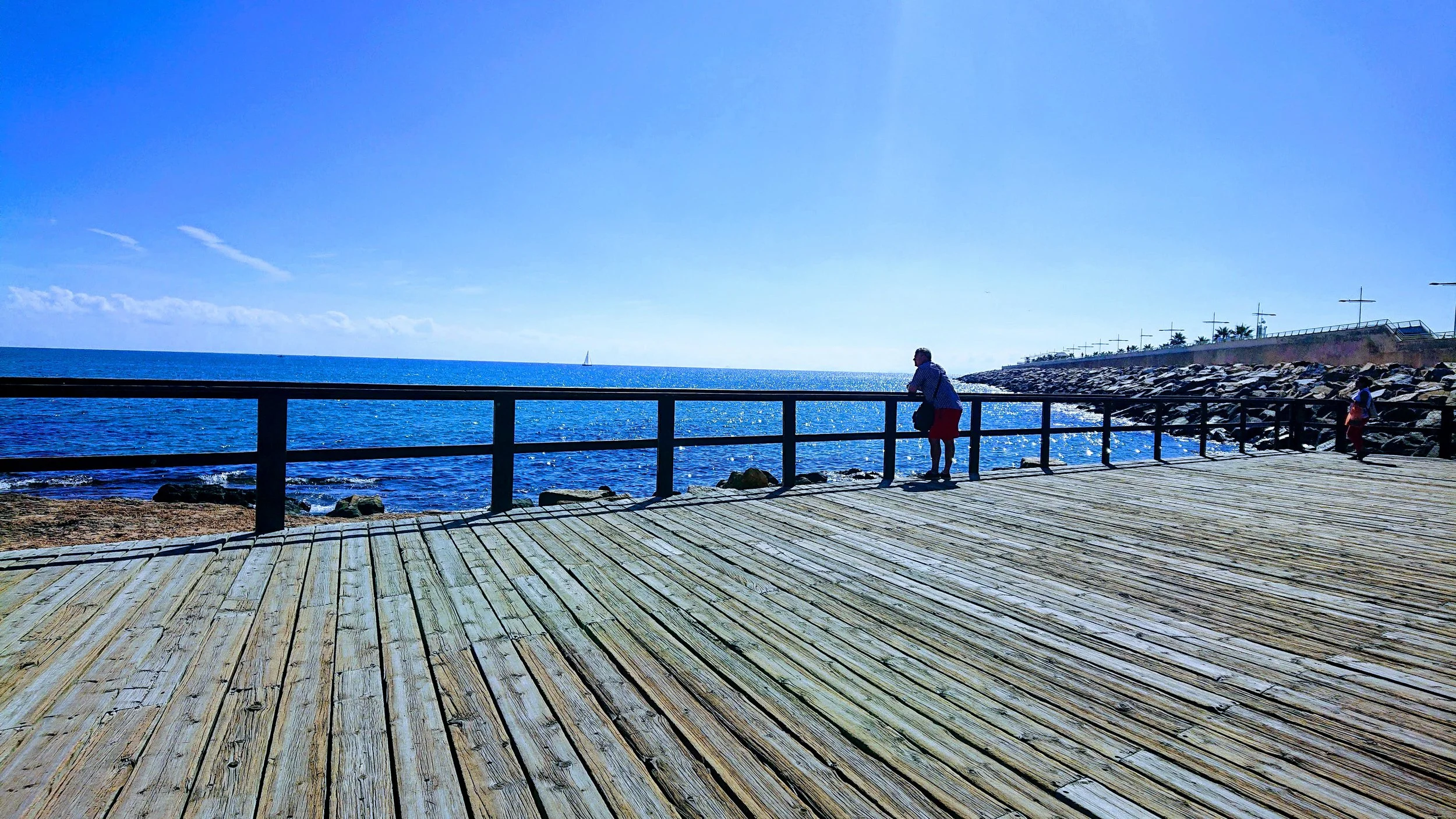 Scenic coastal boardwalk overlooking a bright blue ocean on a sunny day, featuring a person enjoying the sea view from a wooden deck with waves crashing against the rocky shoreline. Perfect serene seaside landscape ideal for travel, tourism, and beac