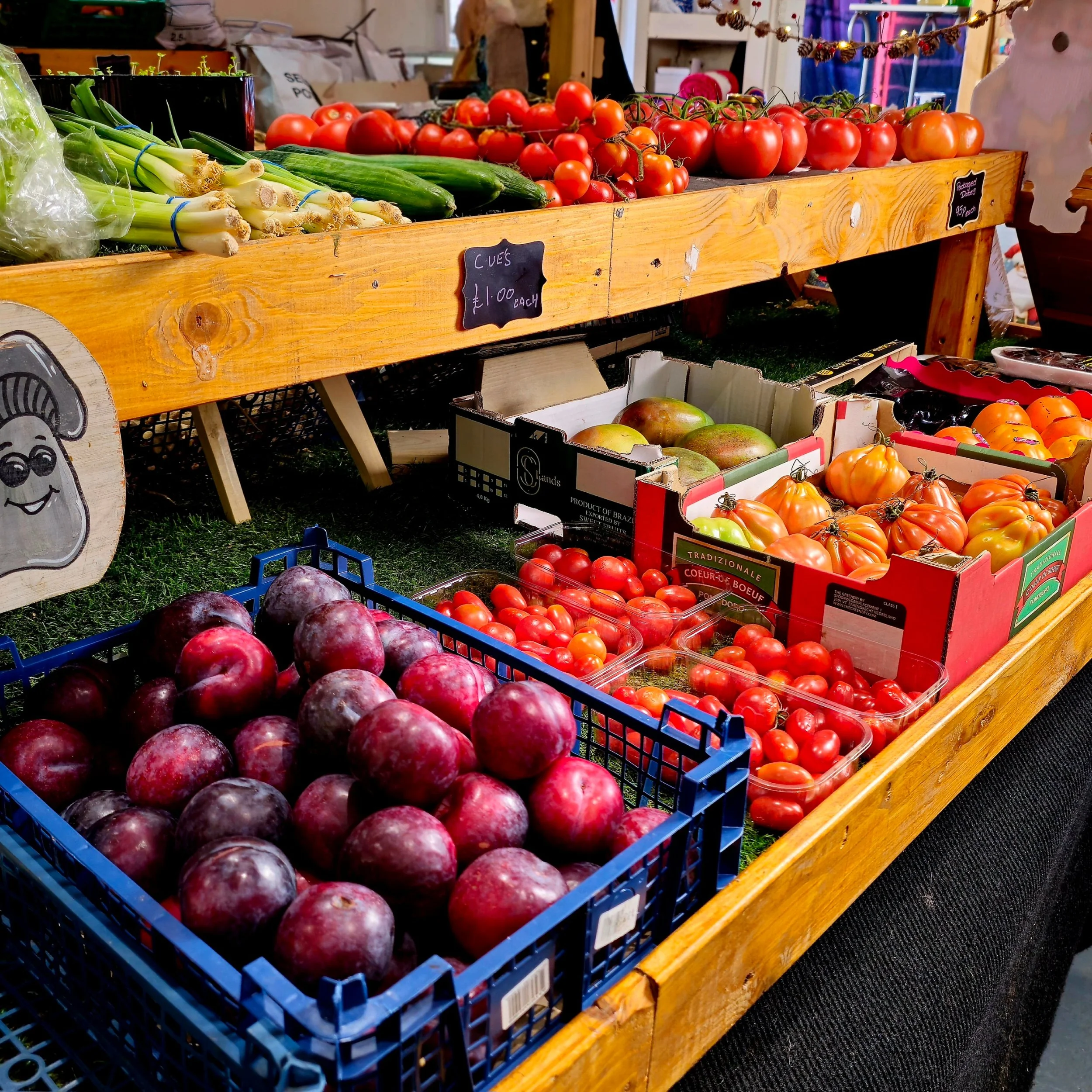 Inside the farm shop, rows of tomatoes, plums, and fresh vegetables create a feast of natural colour. Warm wooden shelving frames the produce, echoing the tones of the earth. Each element—light, texture, and arrangement—tells the story of dedication 