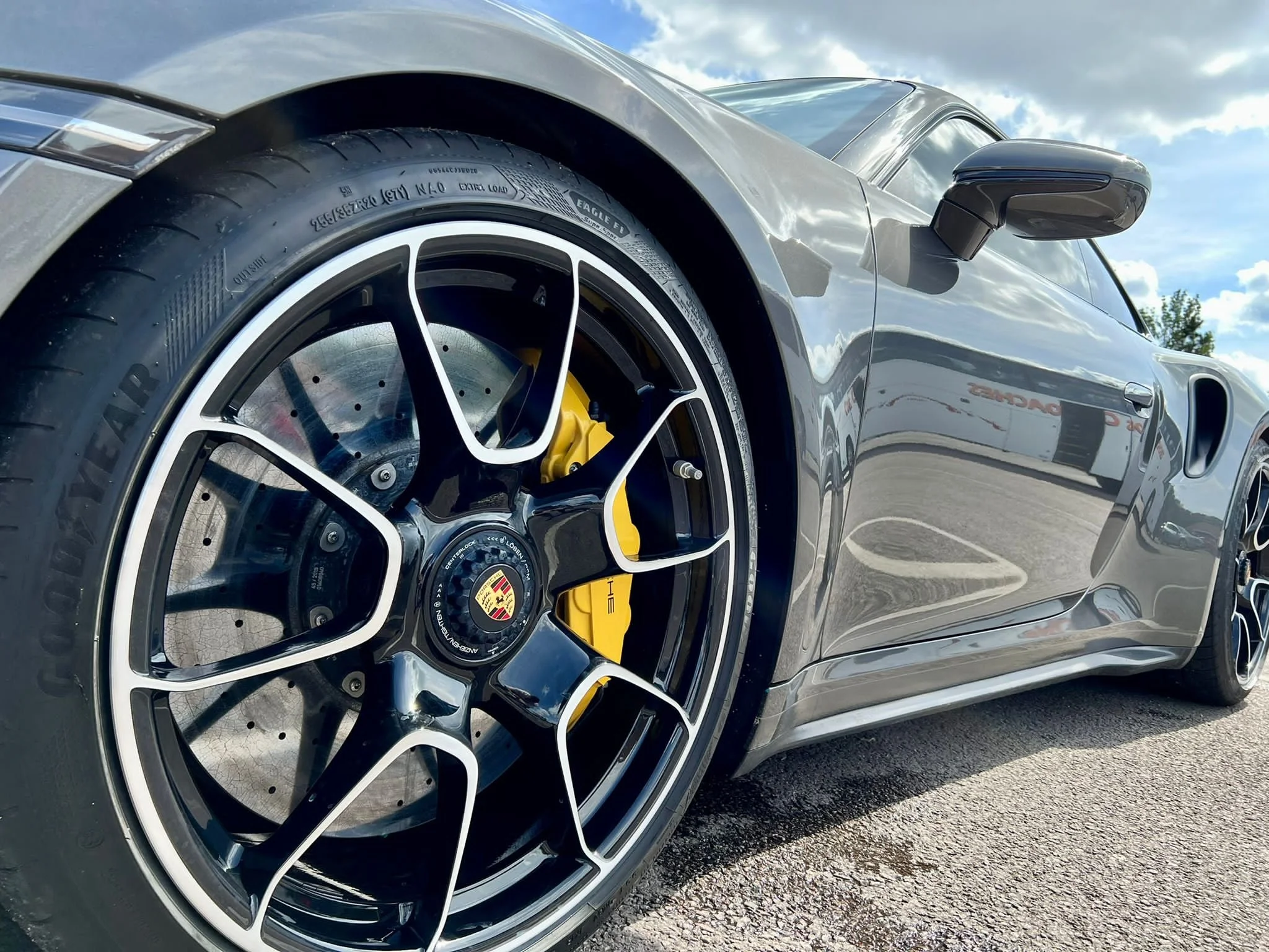 Close-up of the front wheel of a gray Porsche sports car, featuring a black alloy rim with silver accents, yellow brake caliper, and a high-performance tire, parked on an asphalt surface against a partly cloudy sky.
