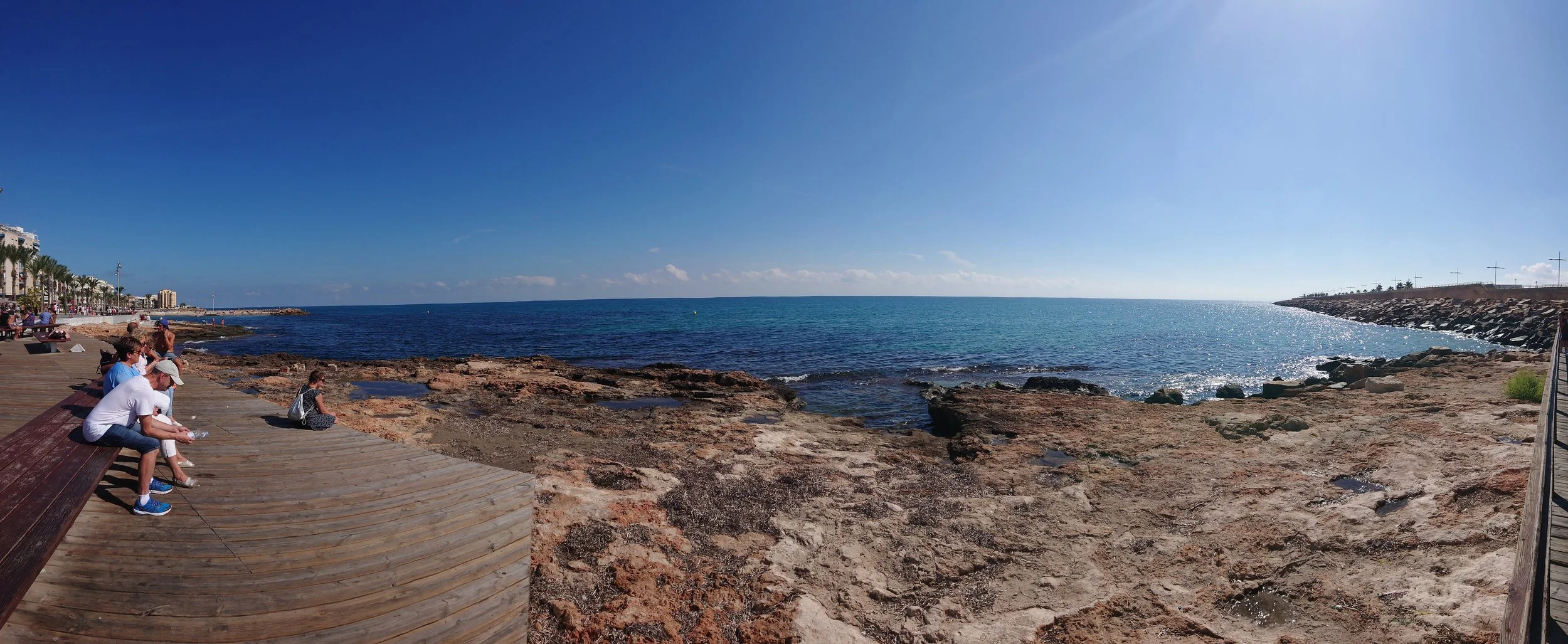 Panoramic seaside boardwalk with visitors relaxing beside a rocky Mediterranean shoreline, overlooking calm turquoise waters and a clear blue sky—capturing a vibrant coastal promenade ideal for travel, tourism, and beach destination websites.