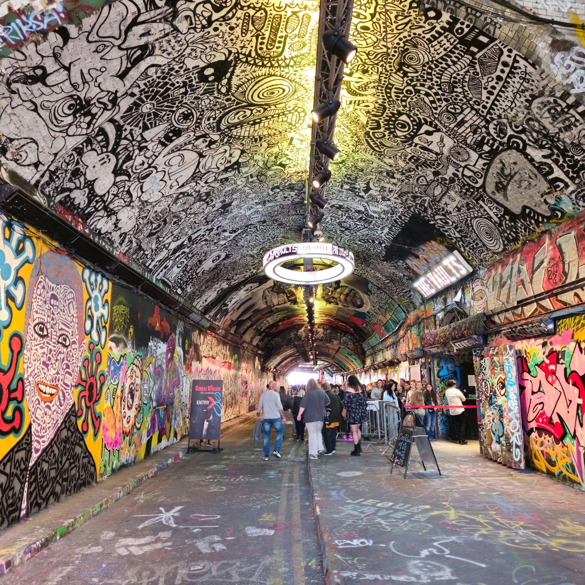 A full immersive street-art tunnel covered from floor to ceiling in detailed murals, tags, and layered spray-paint textures. The photograph captures the energy of an active urban art space, with visitors exploring the tunnel’s vibrant colours and cha