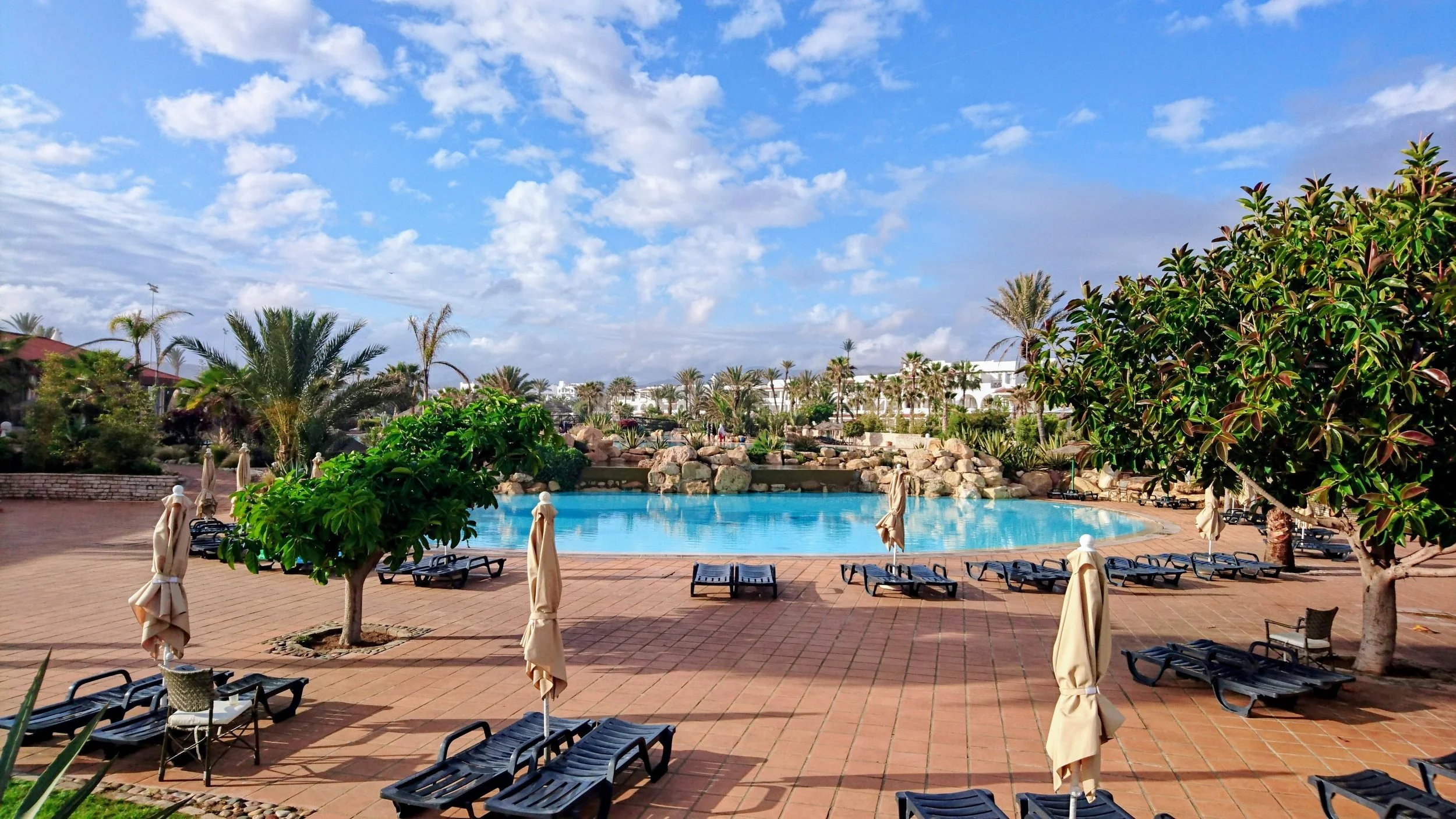 Spacious resort swimming pool surrounded by palm trees, sun loungers, and tropical landscaping under a bright blue sky, creating a relaxing vacation atmosphere ideal for travel, hotel, and luxury getaway websites.