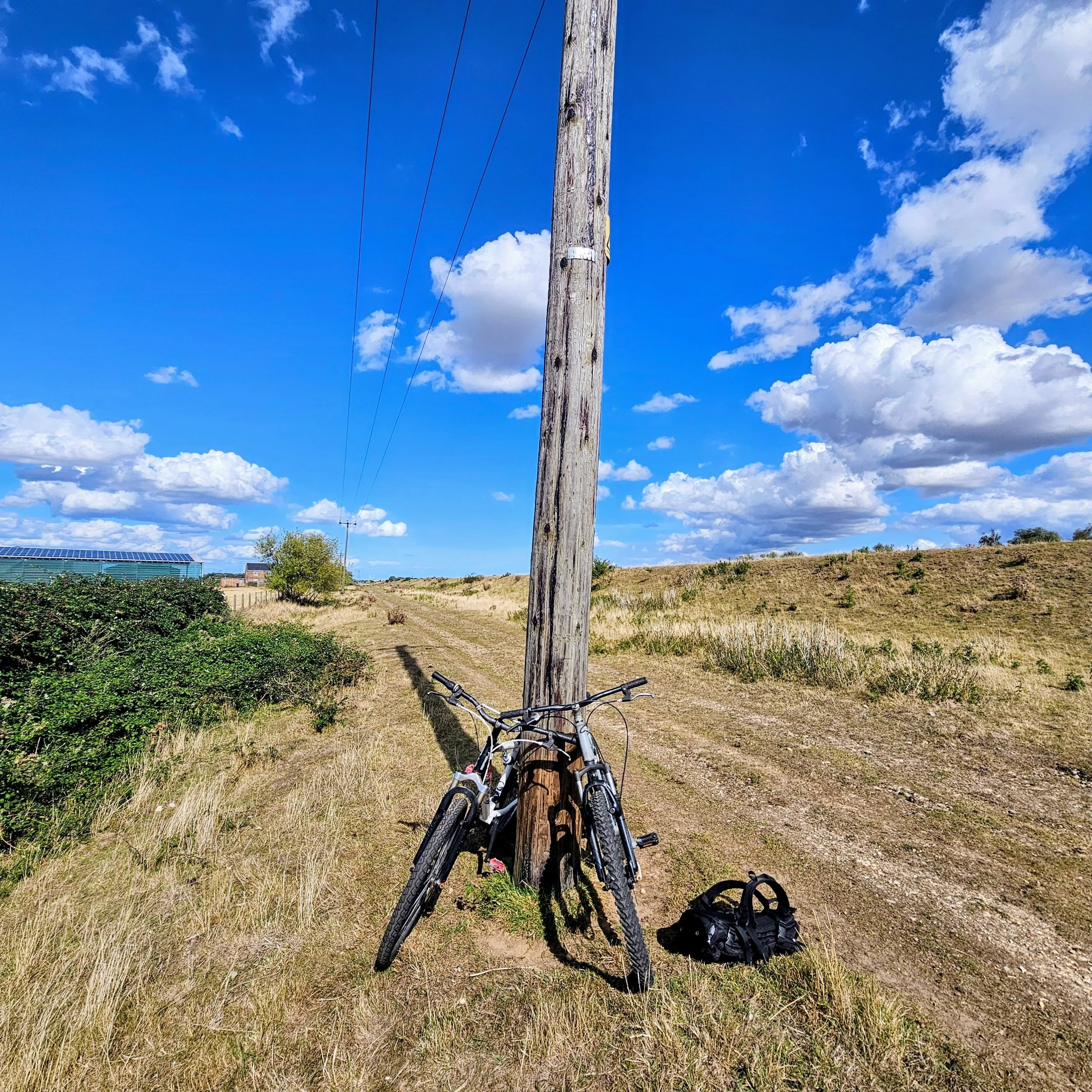Two bicycles leaning against a wooden utility pole on a rural dirt trail under a bright blue sky with scattered clouds, surrounded by dry grassland—scenic countryside cycling and outdoor adventure setting.