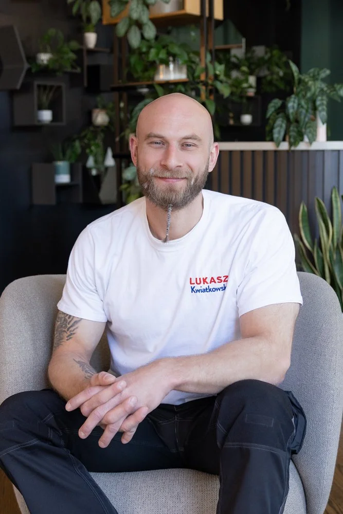 A smiling bald man with a beard sitting on a light-colored armchair in a room with green plants and modern shelves in the background. He is wearing a white T-shirt with red and blue text.