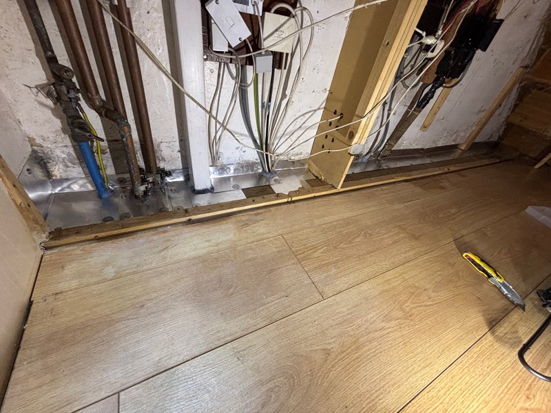Wiring and plumbing visible behind unfinished wall with hardwood flooring being installed.