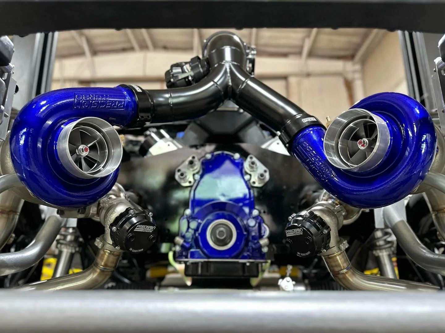 Close-up of a high-performance racing engine with twin turbochargers, blue intake pipes, and metal piping in a garage setting.
