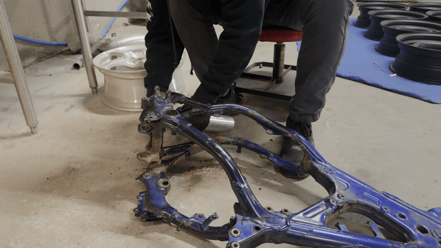 Two people working on a blue motorcycle frame in a workshop, with tools and tires in the background.