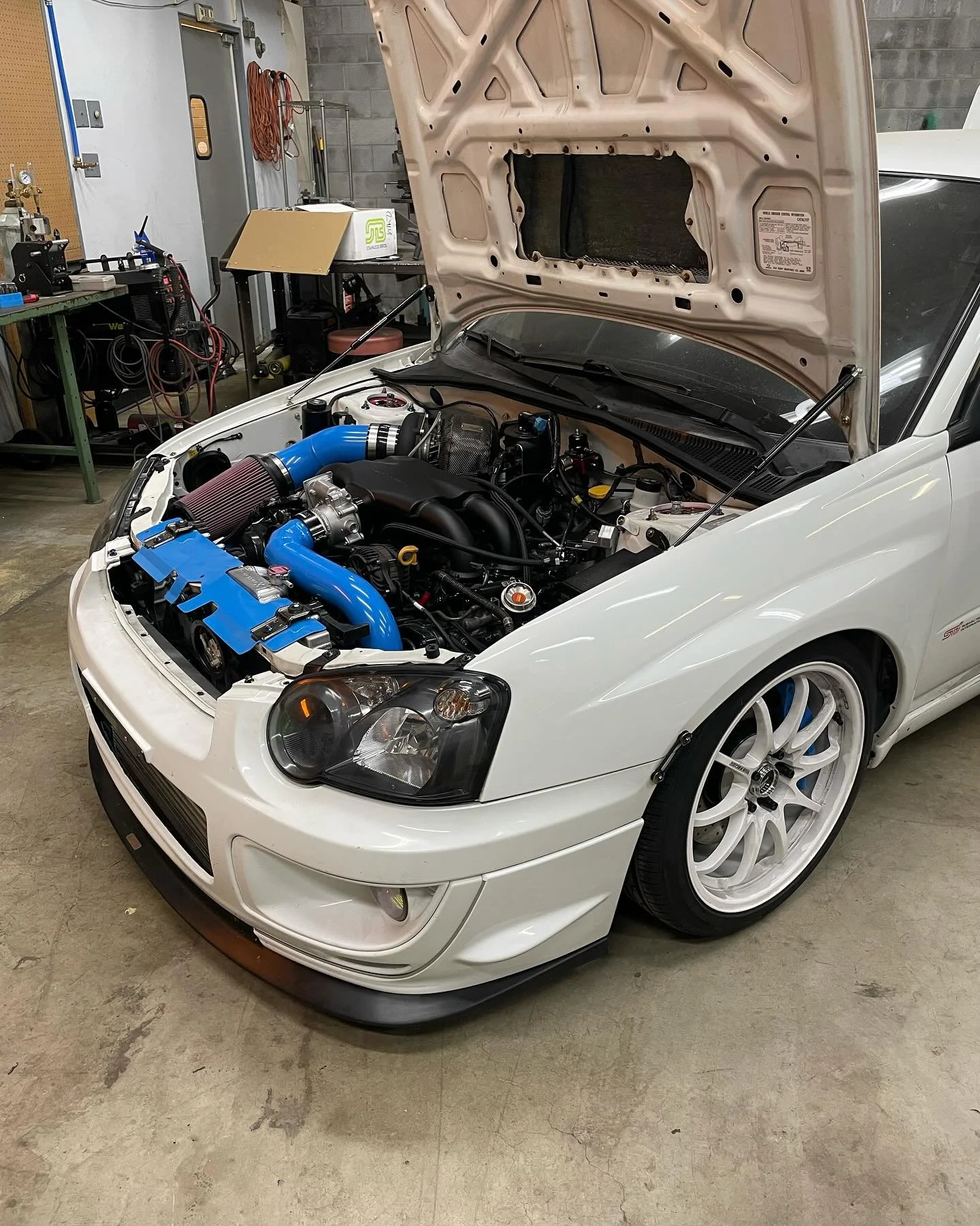 A white sports car with its hood open showing a modified engine bay with blue piping and aftermarket parts inside a garage.