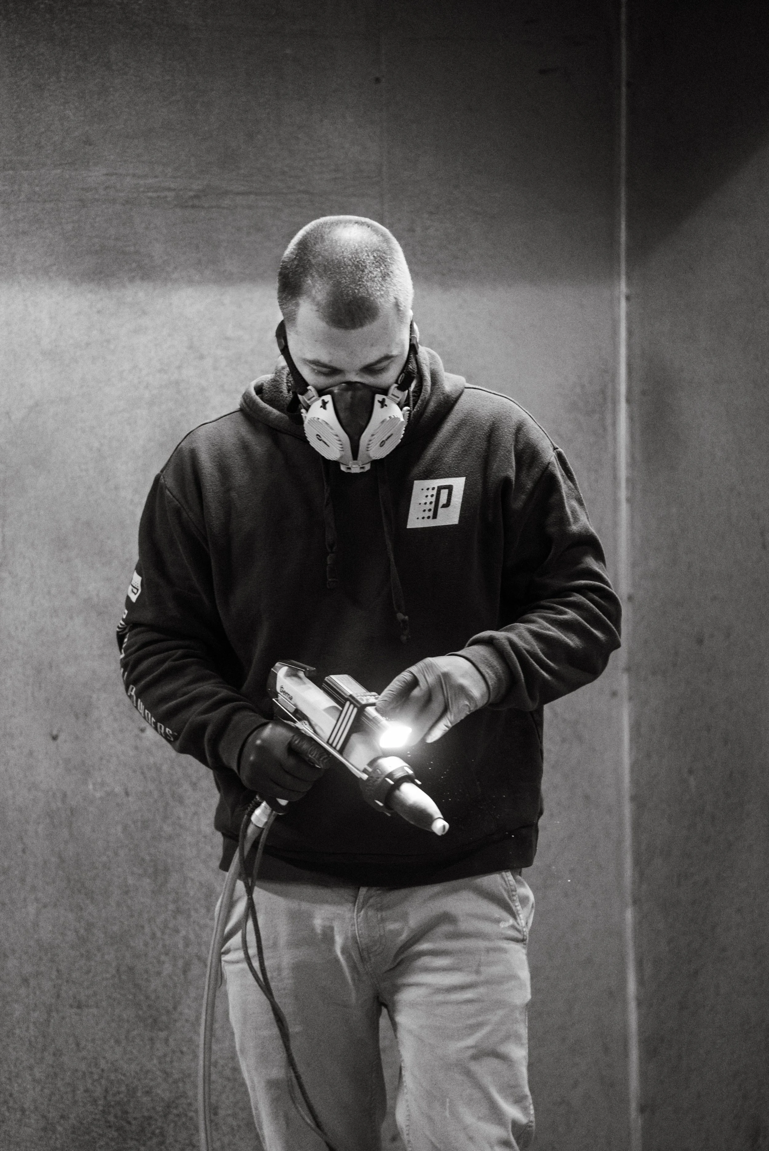 A person wearing a mask, gloves, and a hooded sweatshirt holding a device with a bright light in an indoor setting.