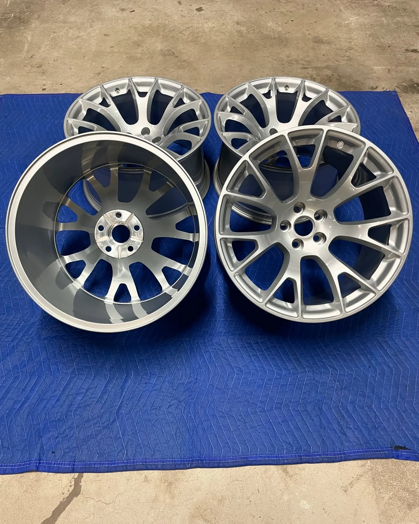 Four silver alloy wheels for a vehicle placed on a blue padded mat on a concrete floor.