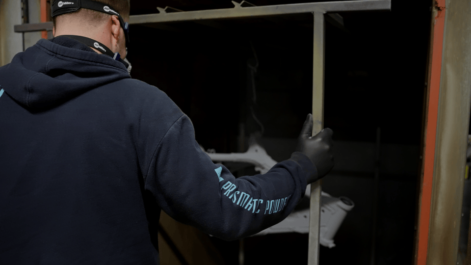 A person wearing a black hoodie and gloves facing away, examining industrial or mechanical parts stored on shelves in a warehouse or workshop.