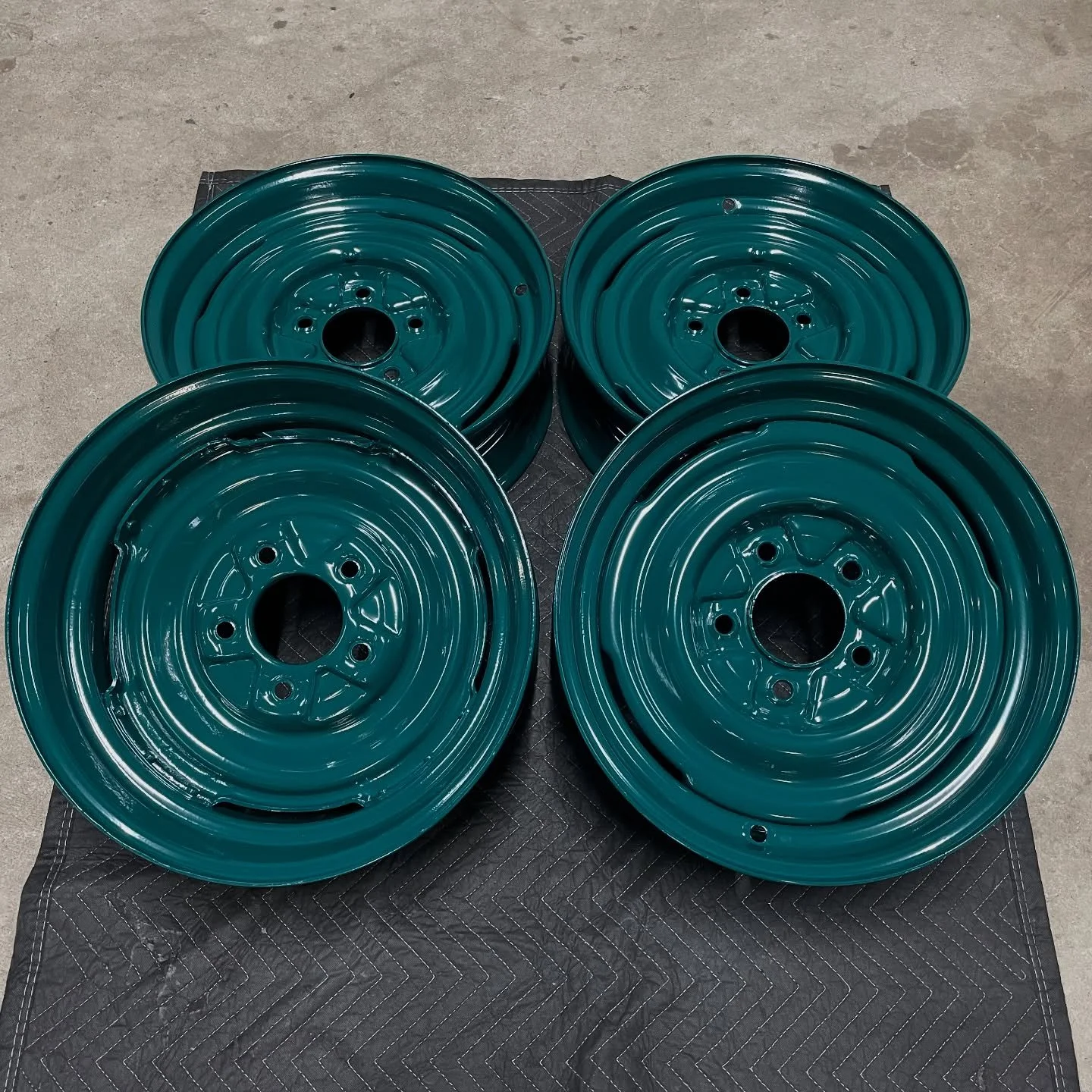 Four teal car wheels arranged in a square pattern on a black quilted mat on a concrete floor.