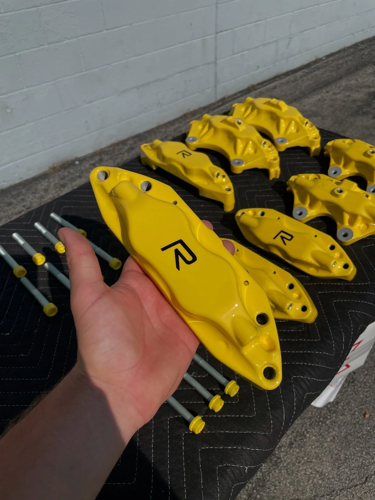 Set of yellow brake calipers and tools on a black quilted surface outside, with a gray brick wall in the background.