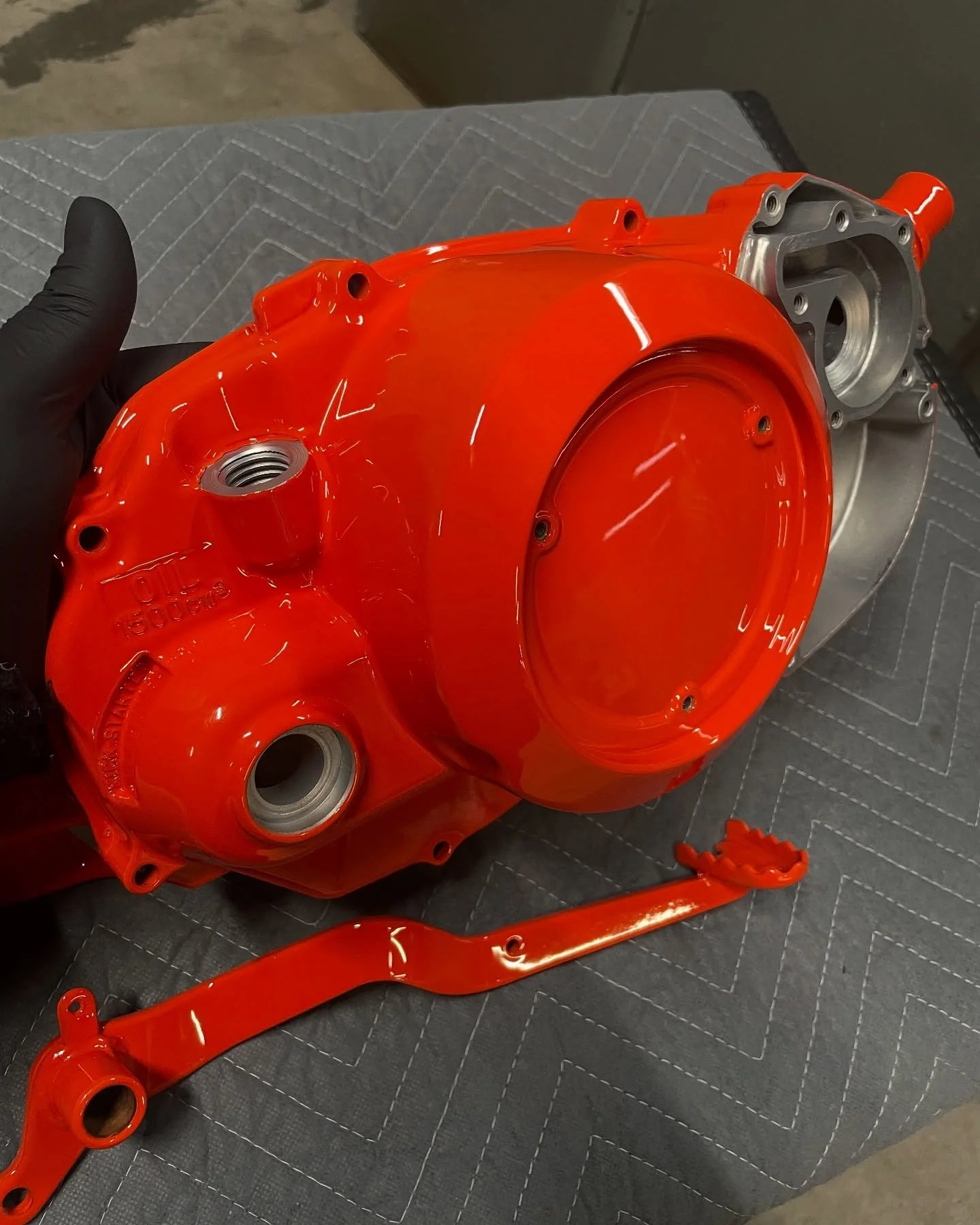 A bright orange mechanical component, likely a motorcycle engine casing, resting on a black quilted surface, with a black gloved hand holding it.