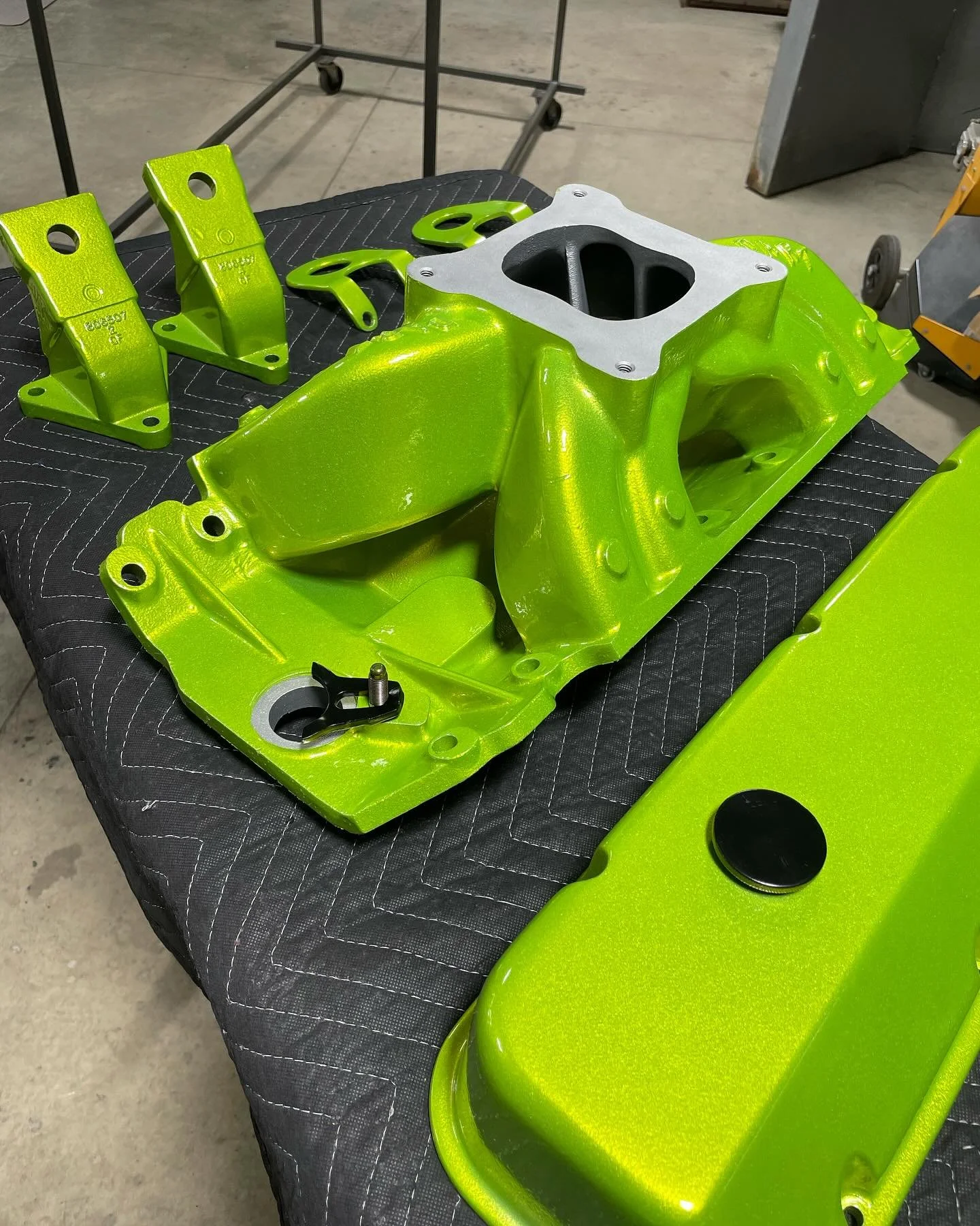 Bright green painted engine parts laid out on a black padded work surface in a workshop, including a valve cover, an intake manifold, and brackets.