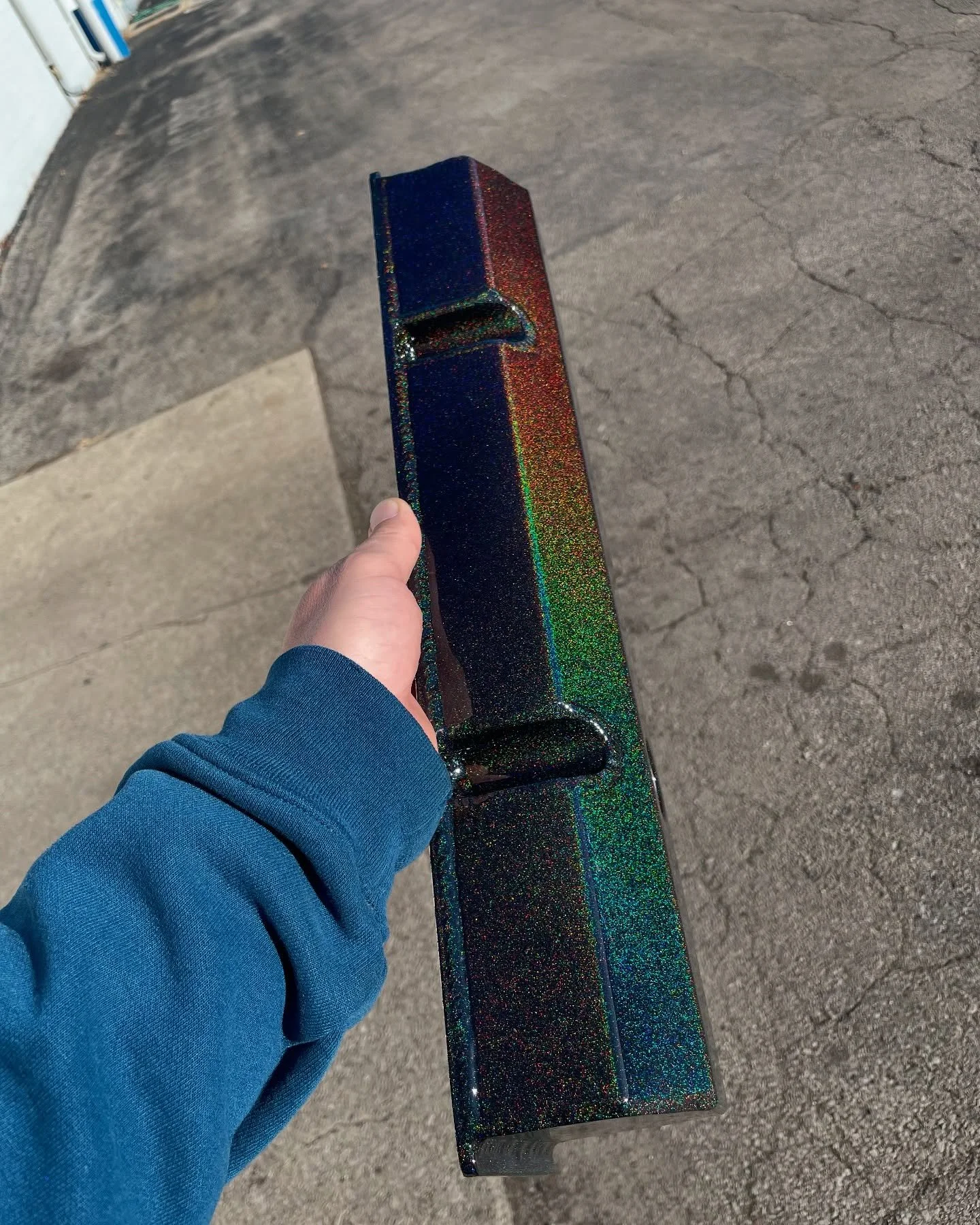 A person wearing a blue hoodie holding a sparkly, iridescent dark-colored box or object outdoors on a concrete surface.