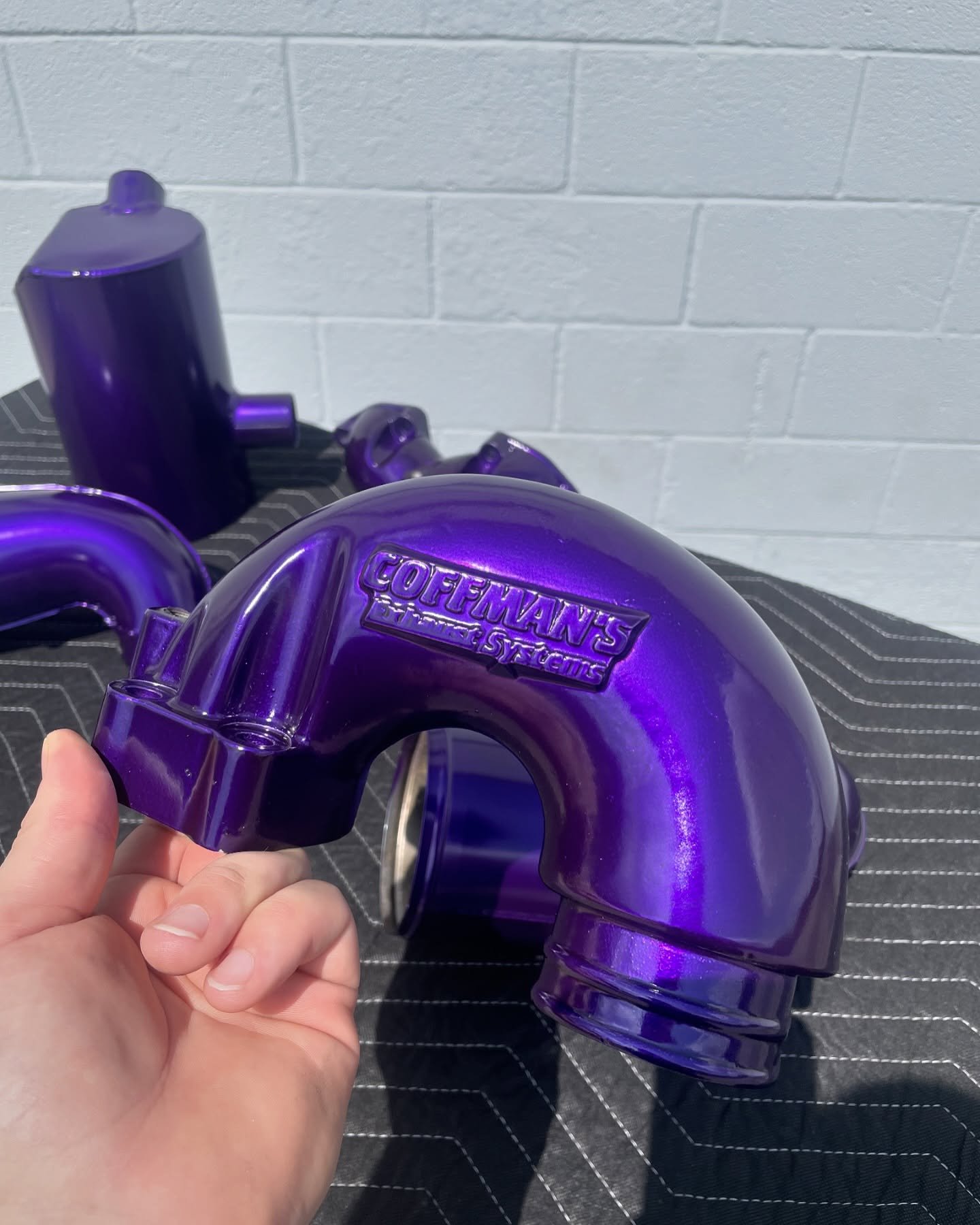 A purple metallic exhaust pipe with the logo 'Coffmans' on it, held by a person's hand, resting on a black surface with a white brick wall in the background.