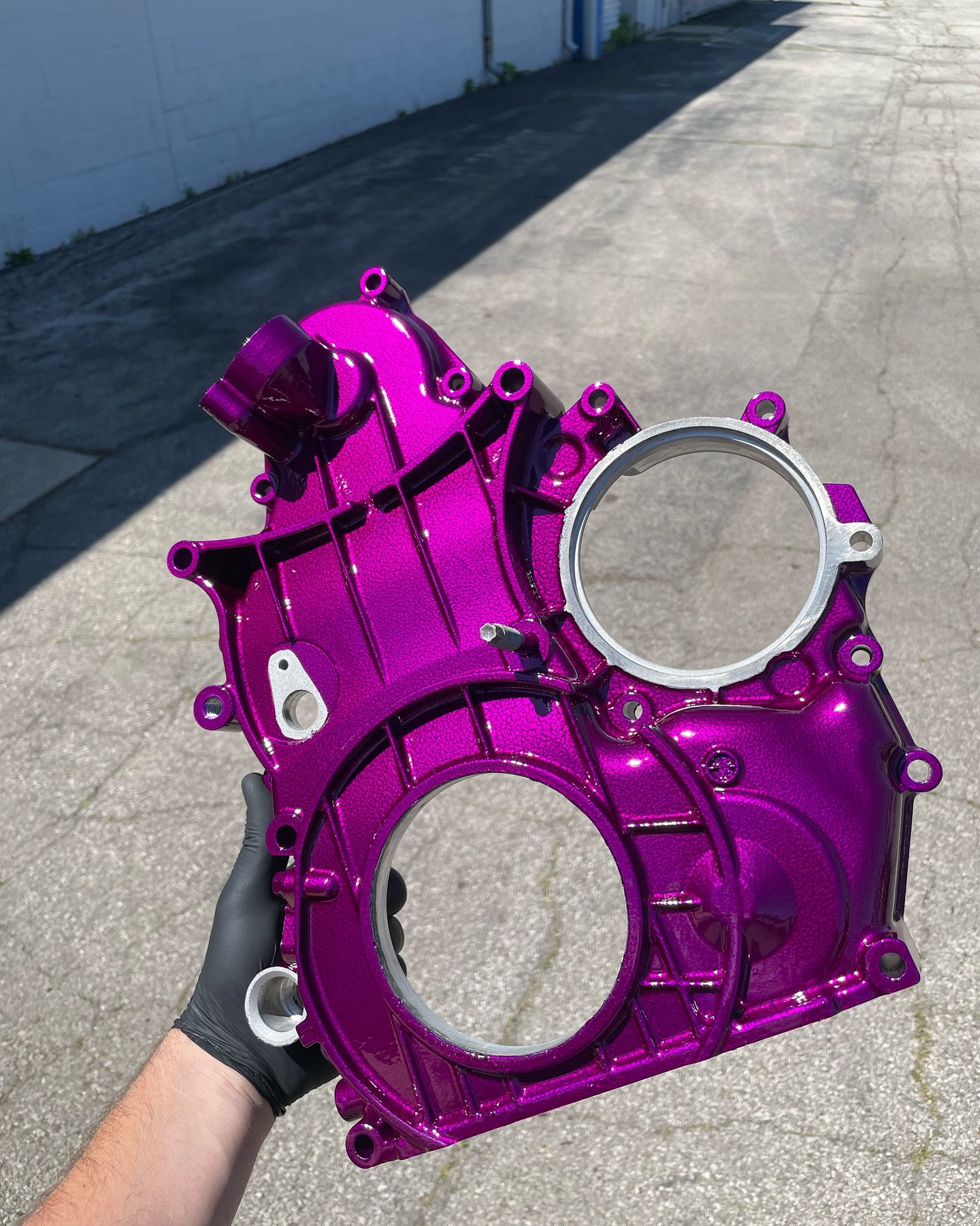 Person holding a purple metallic engine cover with two large circular openings, outdoors on concrete ground.