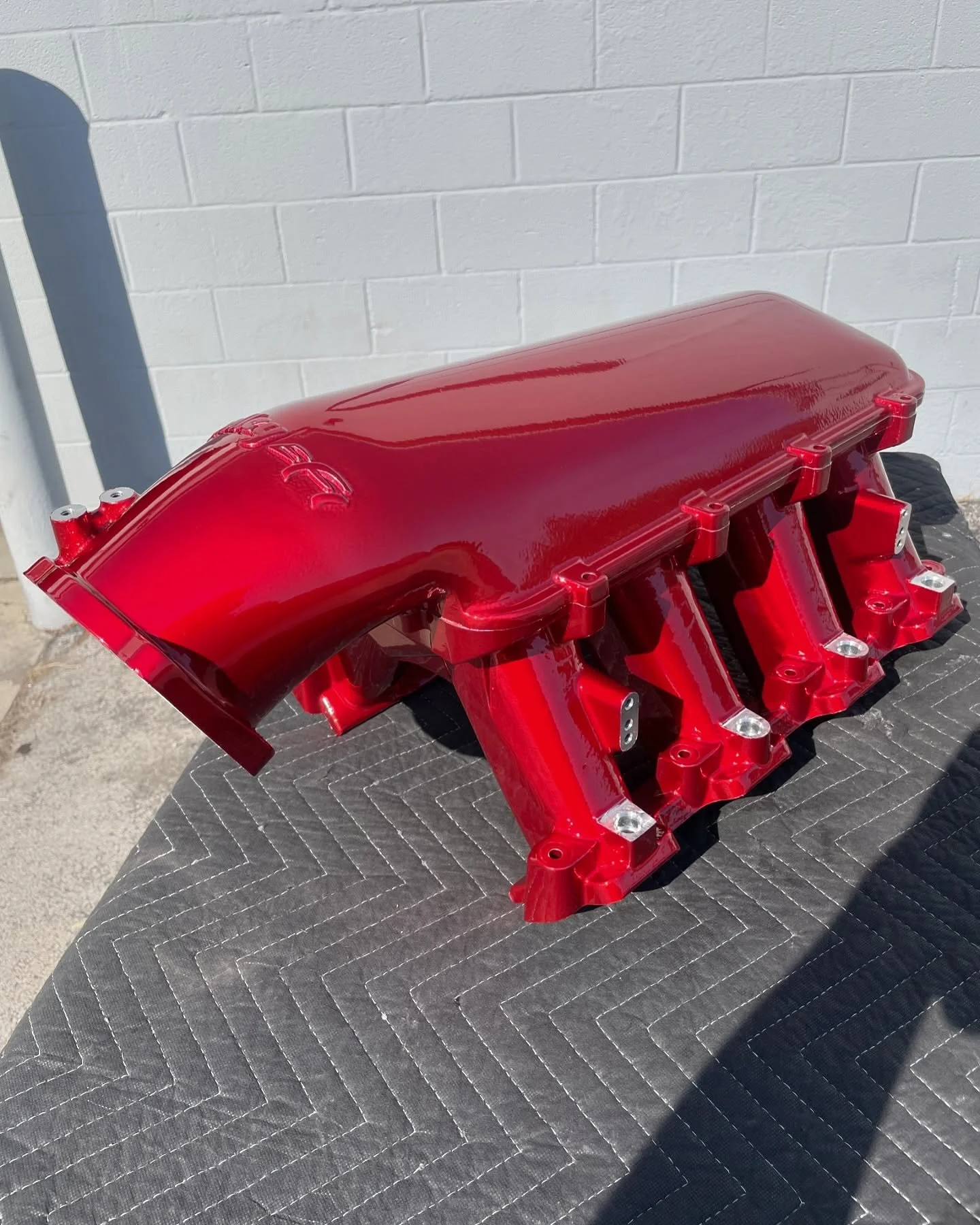 A red painted automotive engine intake manifold on a black quilted surface outdoors.
