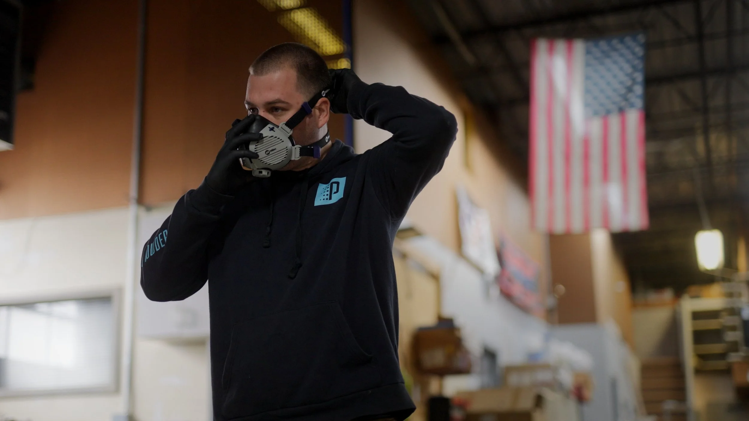 A person wearing a black hoodie and gloves, adjusting a protective face mask inside a large indoor space with American flags hanging from the ceiling.