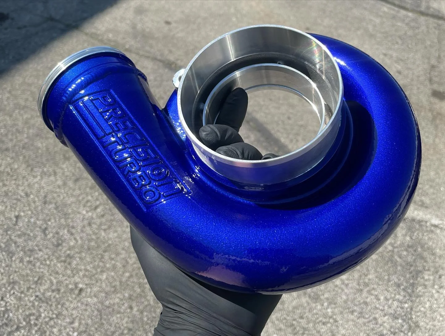 Hand wearing a black glove holding a blue turbo charger with the brand name 'Precision Turbo' embossed on it, outside on a sunny day.