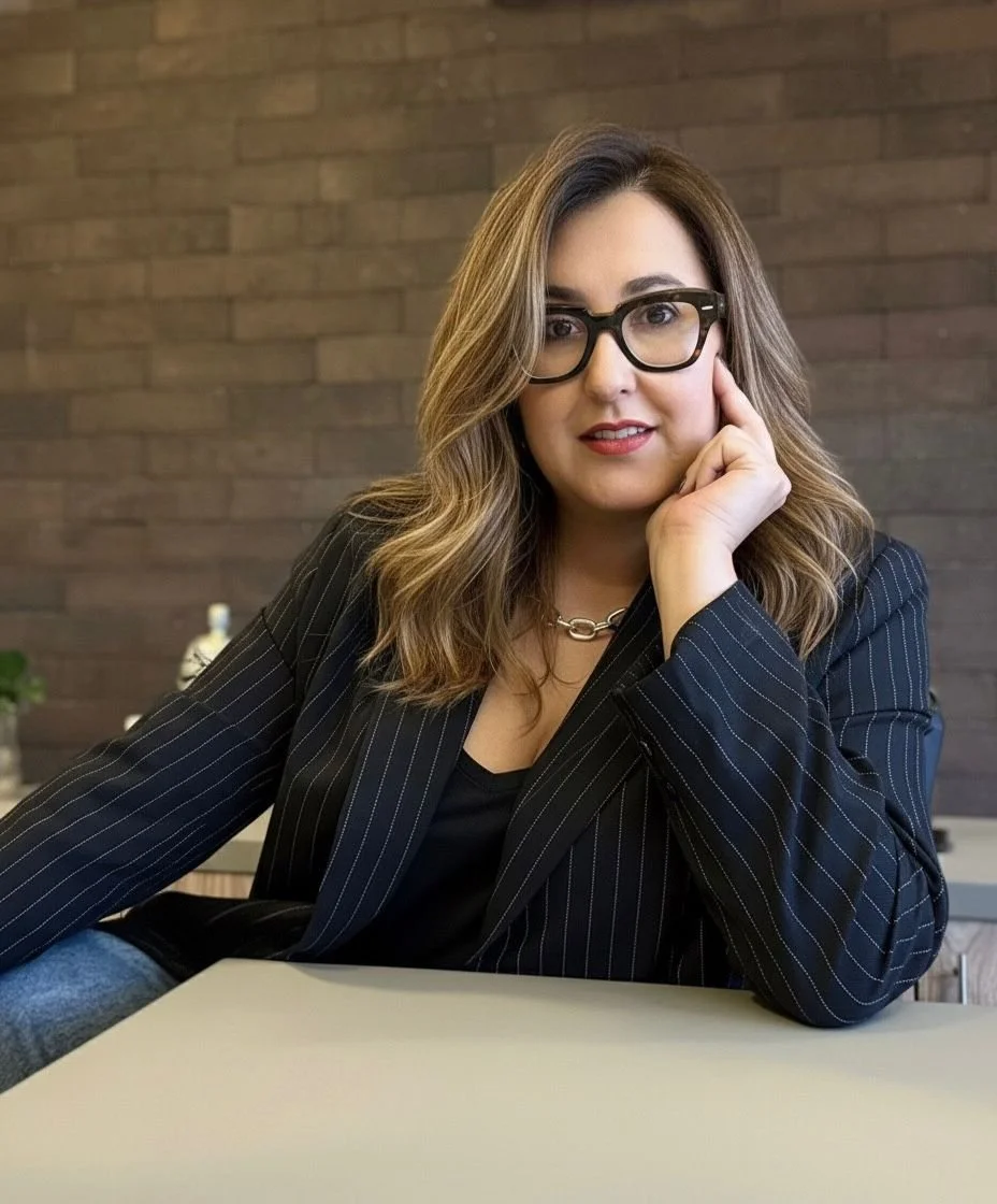 Mulher com cabelo castanho, óculos pretos, vestido ou blazer preto com riscas, sentada à mesa com um fundo de parede de tijolos, sorrindo levemente.