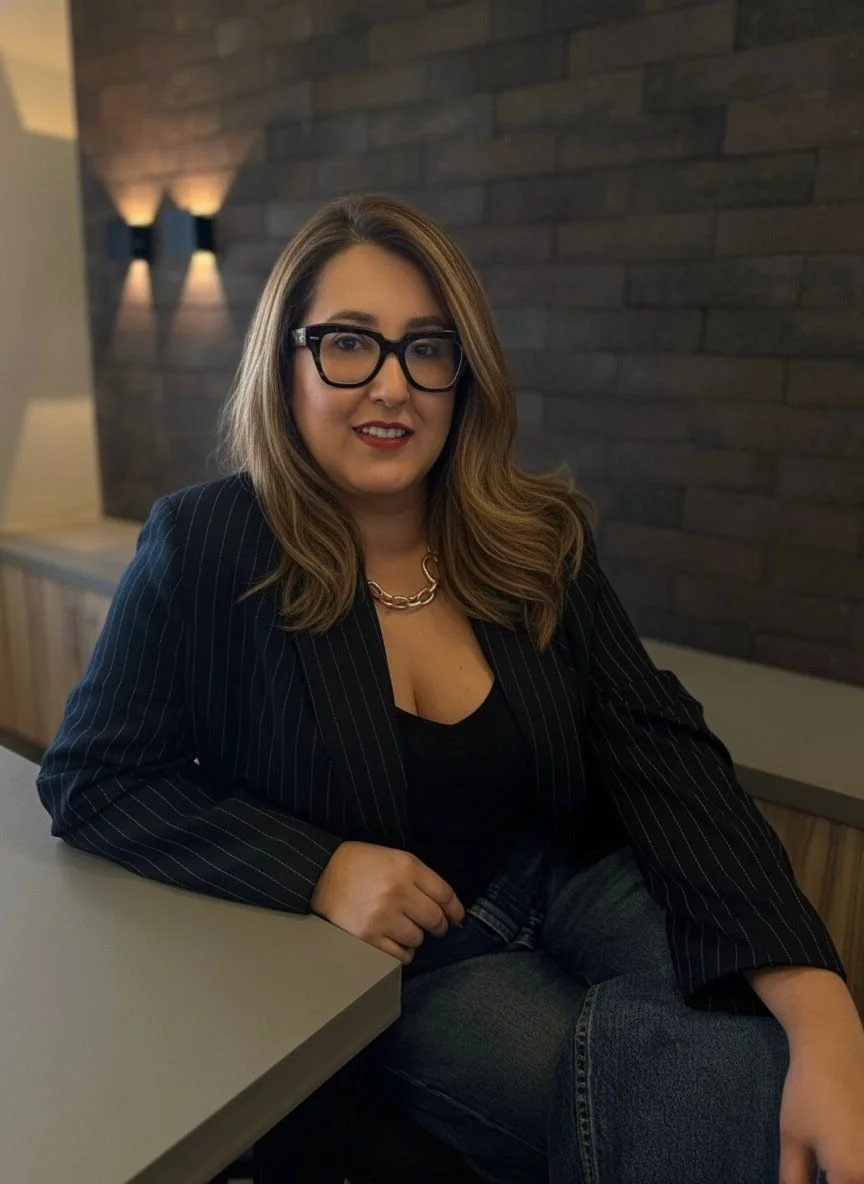 Mulher de cabelos castanhos e lisos, usando óculos grandes e uma blusa preta com blazer riscado, sentada em um ambiente com parede de tijolos ao fundo.