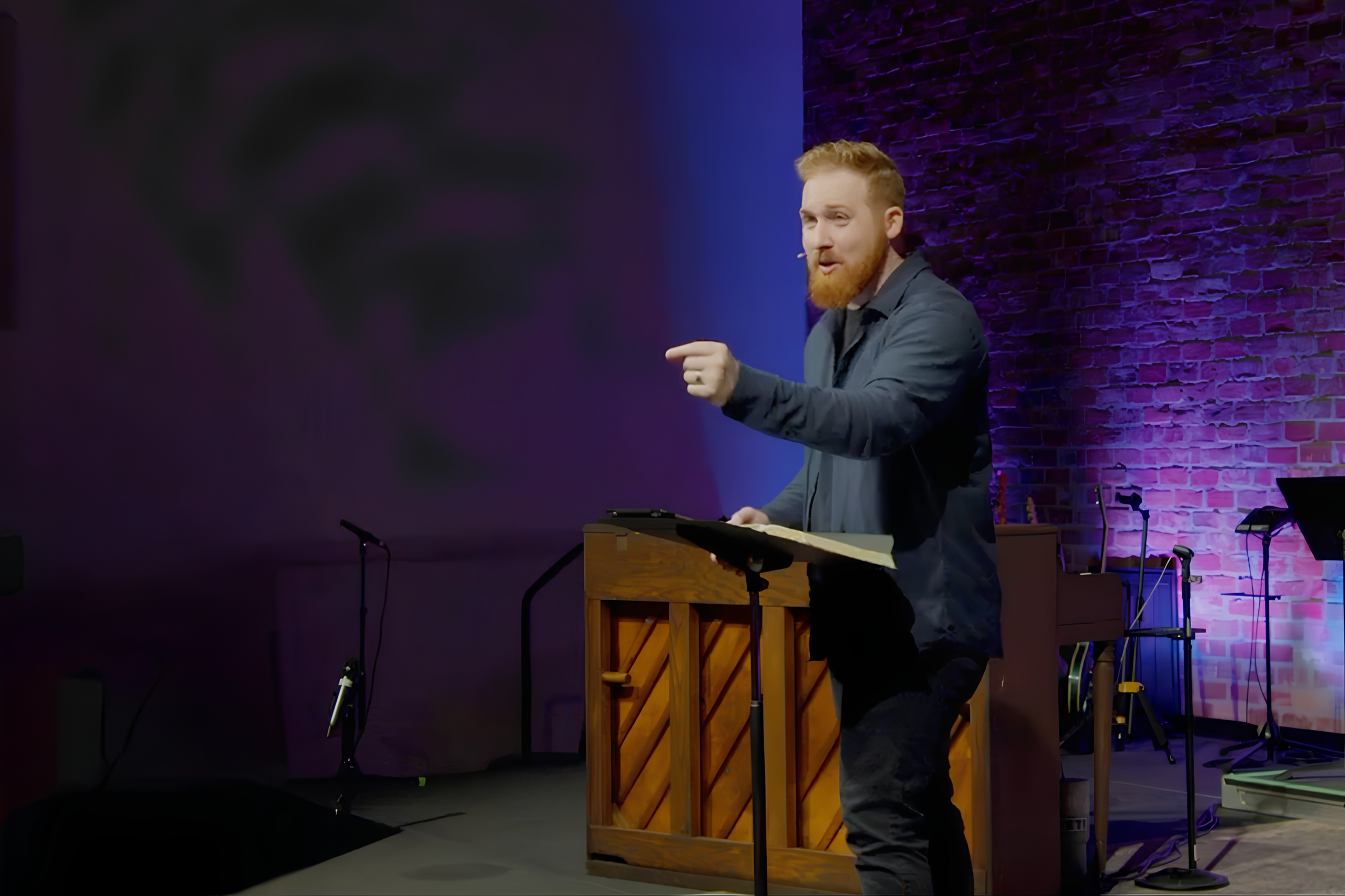 A man preaching with a beard and red hair speaking at a podium on stage with a brick wall background illuminated by purple and blue lights. Risen Church Eindhoven.