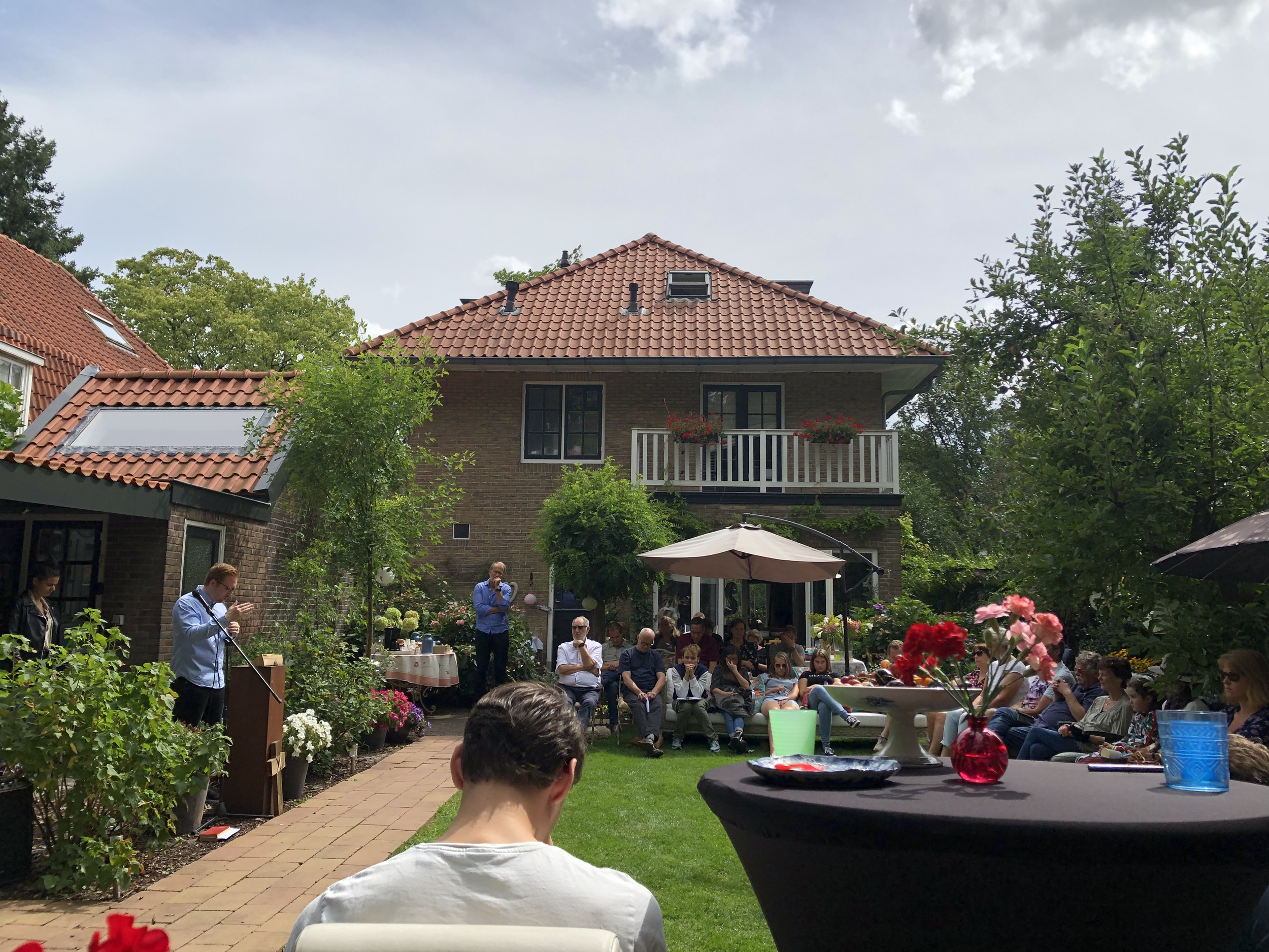 A backyard gathering with people sitting and listening to a speaker, decorated with flowers, umbrellas, and a table, with a two-story brick house with a balcony in the background.