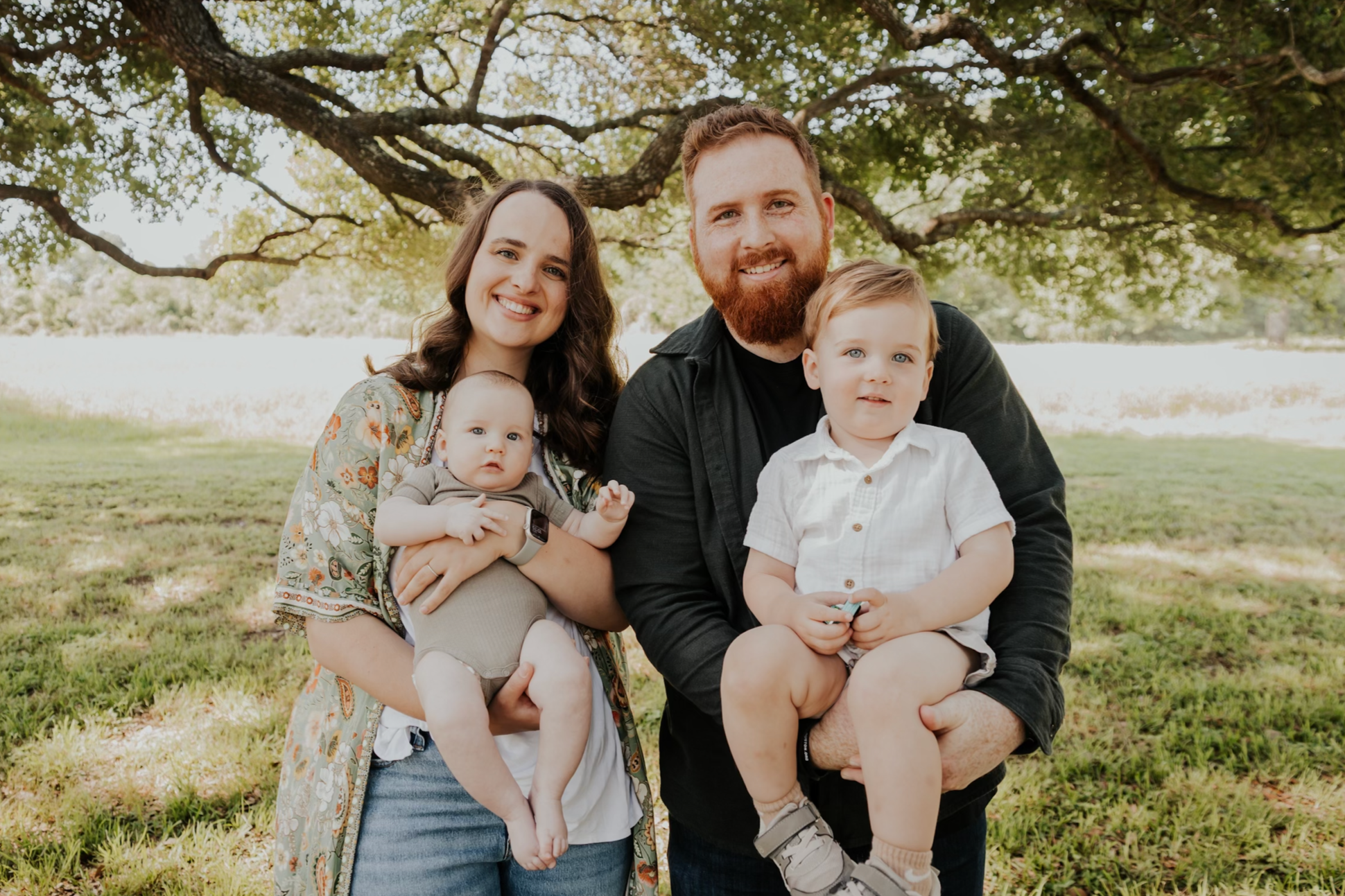 A family of four smiling outdoors under a large tree, with a woman holding a baby and a man holding a young boy, all on grass with sunlight filtering through leaves.