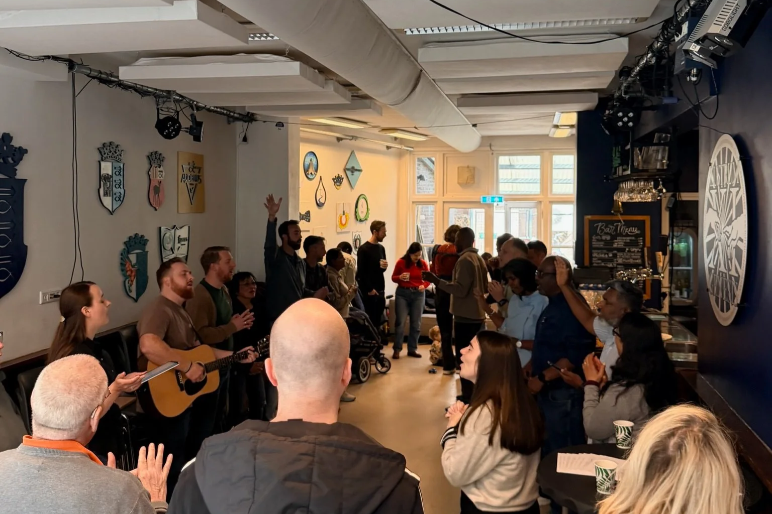 Group of people gathered in a cafe or community space, some are standing, some sitting, with a person playing guitar and others singing or clapping. The room has decorative wall art, large windows.. Risen Church Eindhoven.