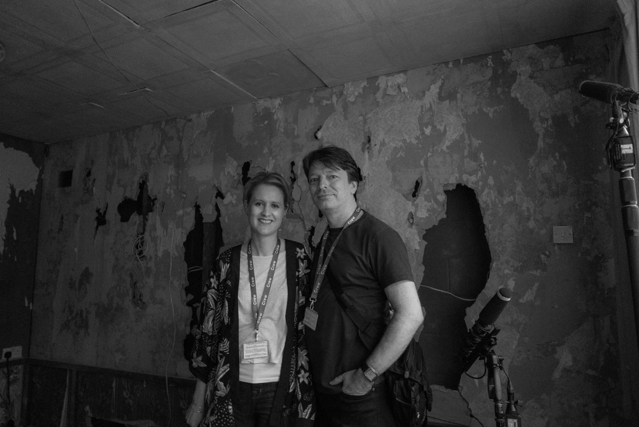 Felicity Evans and Toby Venables, screenwriters and screenwriting consultants, standing in front of a damaged wall with peeling paint, wearing lanyards with badges