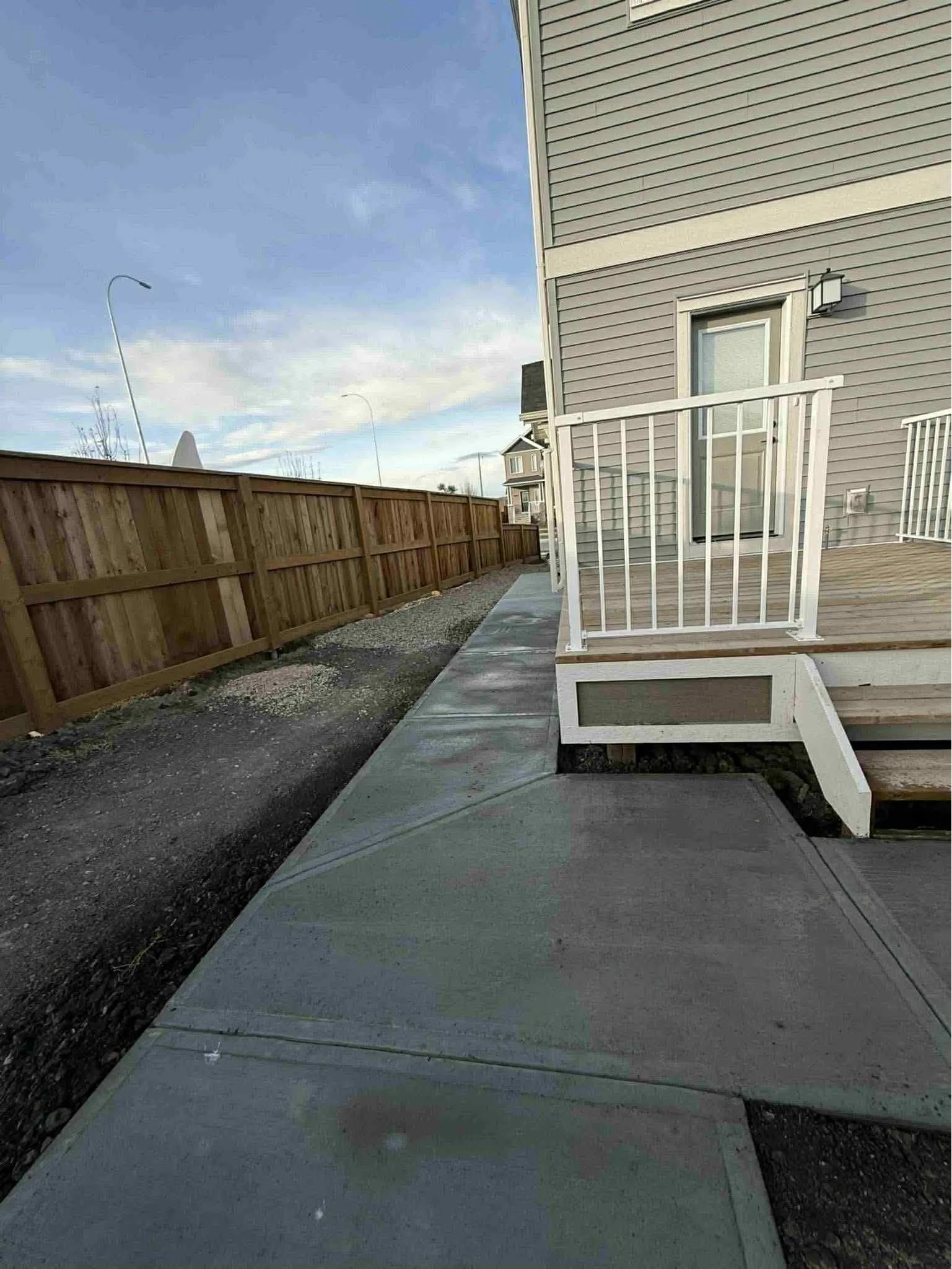 Side view of a residential house's back porch with white railing and steps, surrounding a concrete patio and walkway, fenced backyard, neighboring houses, and a partly cloudy sky.