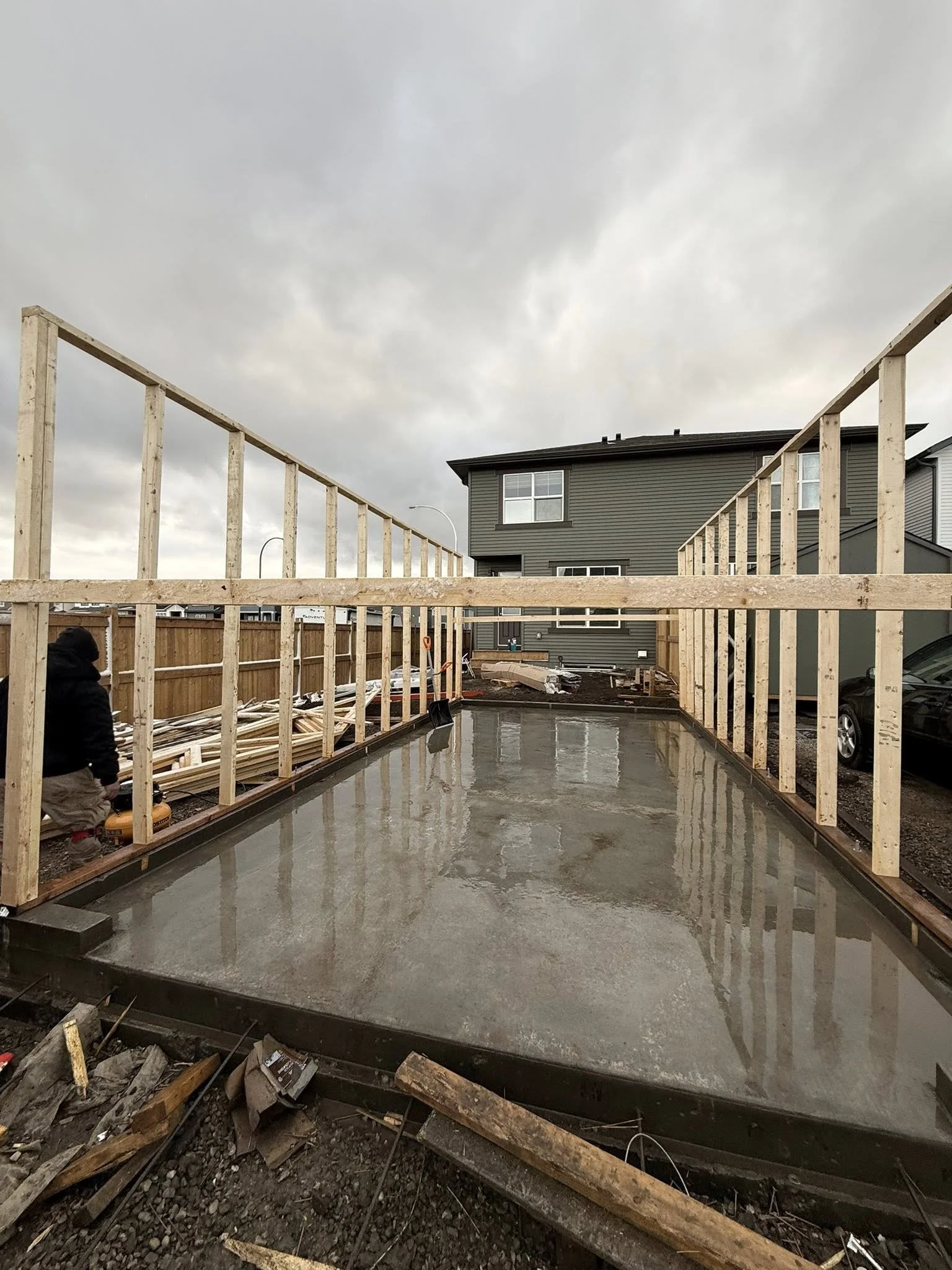 Construction site with a concrete foundation and wooden framing for a building, with a residential house in the background under cloudy sky.