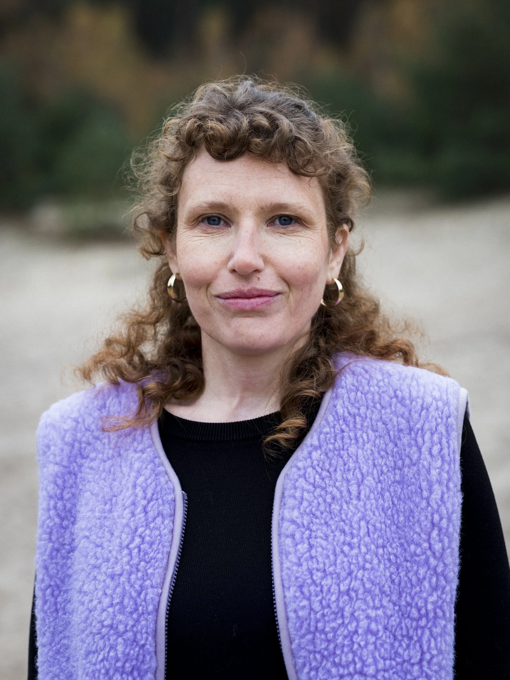 A woman with curly red hair, blue eyes, and wearing gold hoop earrings, standing outdoors in front of a blurred green and brown background, wearing a black shirt and a lavender fleece vest.