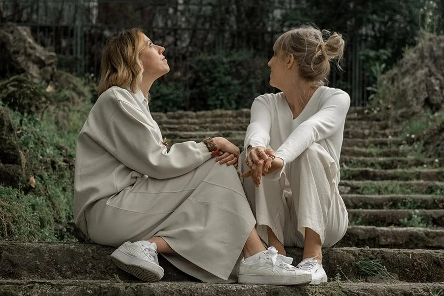 Due donne sedute su dei gradini di pietra in un ambiente naturale, che si tengono per mano e si guardano affettuosamente.