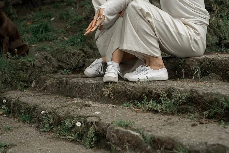 Persona seduta su gradini in un ambiente naturale, con scarpe da ginnastica bianche e abbigliamento beige, accanto a un animale, forse un cane o un altro mammifero, e con alcune piante e fiori selvatici intorno.