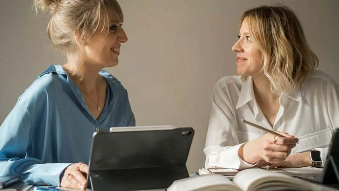 Due donne in un incontro di lavoro o consulenza, una con camicia bianca e l'altra con camicetta azzurra, sedute a una tavola con computer e libri.