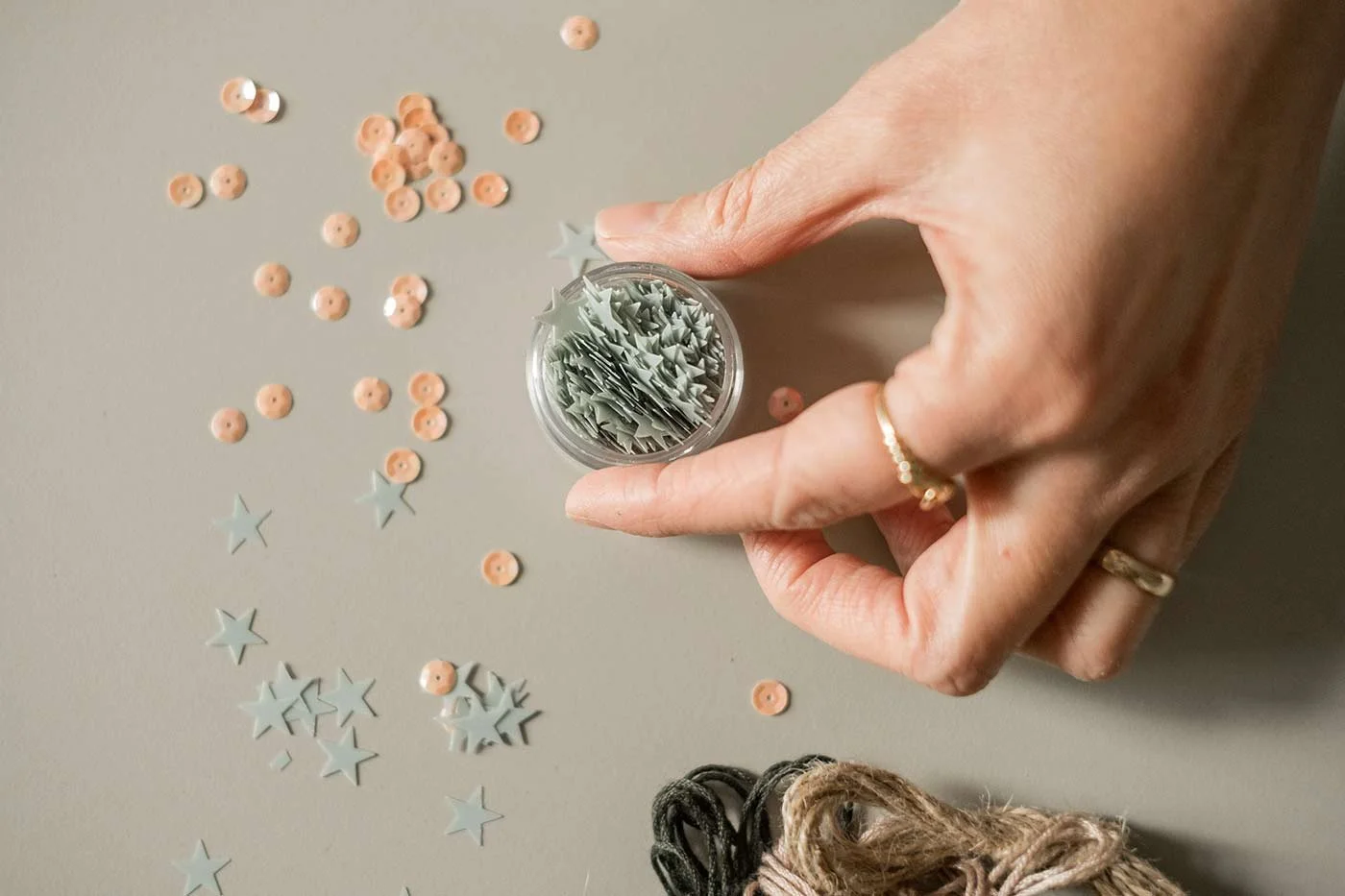 Dita una mano che tiene un contenitore con stelle di carta grigie e piccoli pulsanti rosa, su una superficie chiara, con alcune stelle e pulsanti sparsi.