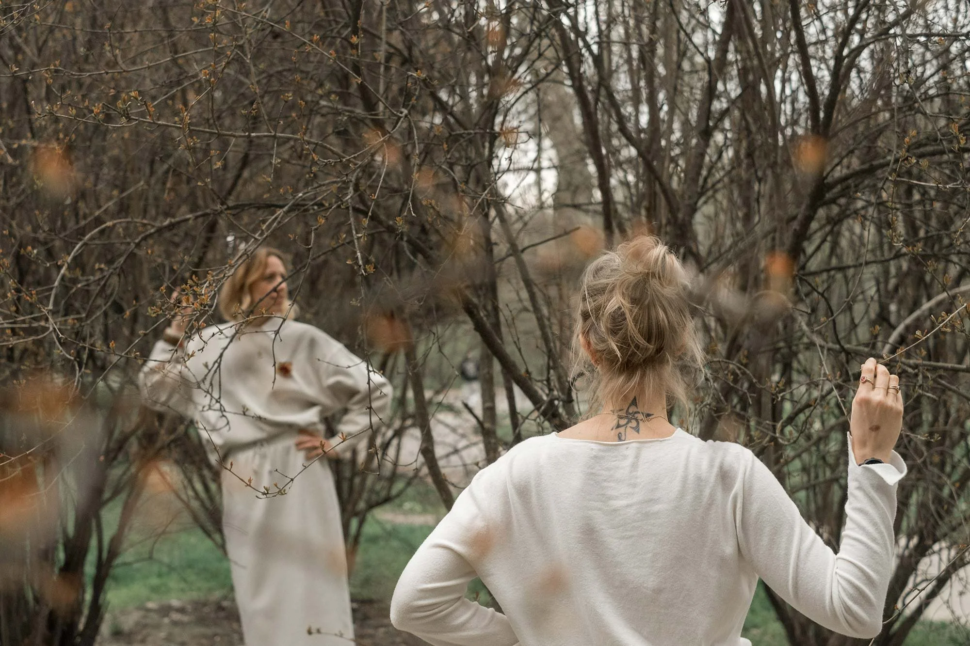 Due donne in un parco con alberi spogli durante l'autunno o l'inverno, una di spalle con i capelli raccolti, che si rivolge verso l'altra donna con occhiali e vestiti chiari, in posa rilassata.