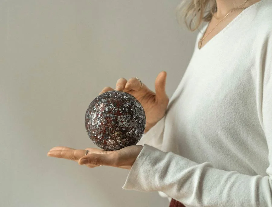 Una donna tiene una sfera decorativa di mosaico metallico con colori scuri e riflessi sulla mano aperta.