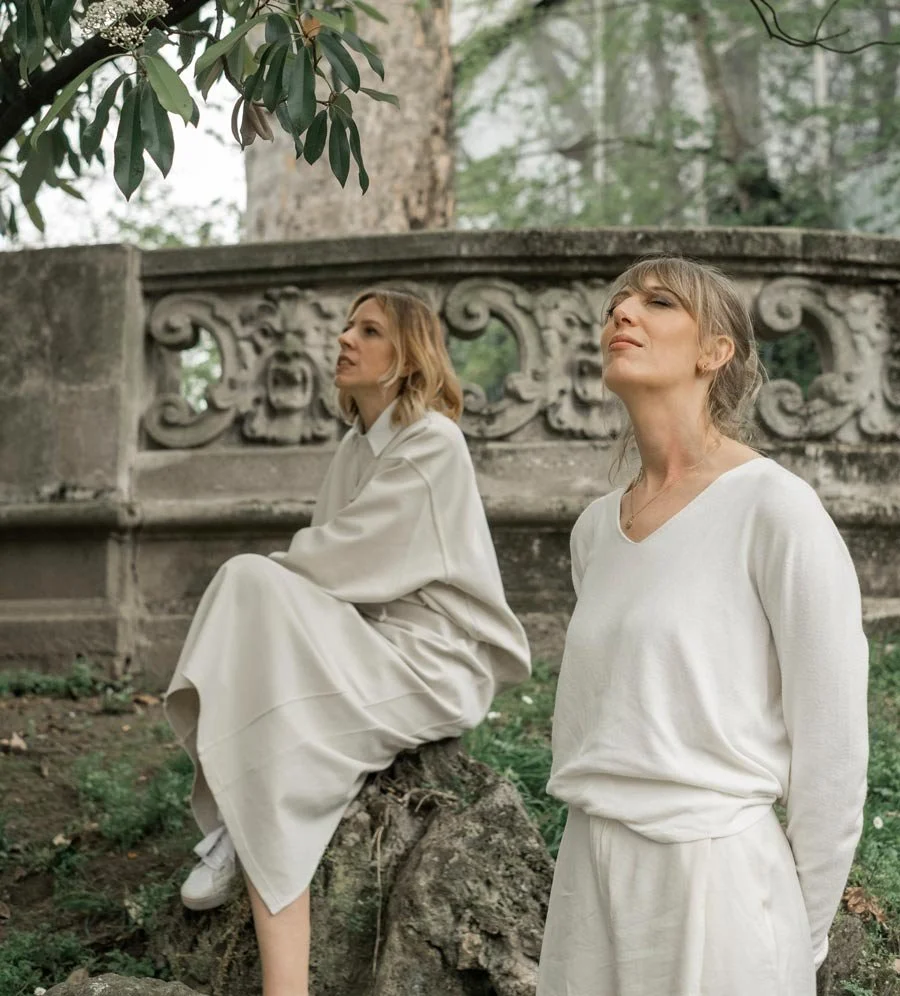 Due donne con abiti chiari si trovano in un giardino accanto a una struttura con un motivo decorativo in pietra. Una delle donne si siede su un tronco di albero, l'altra sta in piedi, entrambe sembrano contemplative o serene.