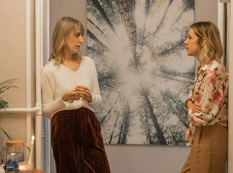 Due donne stanno conversando in una stanza decorata con un quadro di alberi visto dal basso che mostra le chiome delle piante contro il cielo.