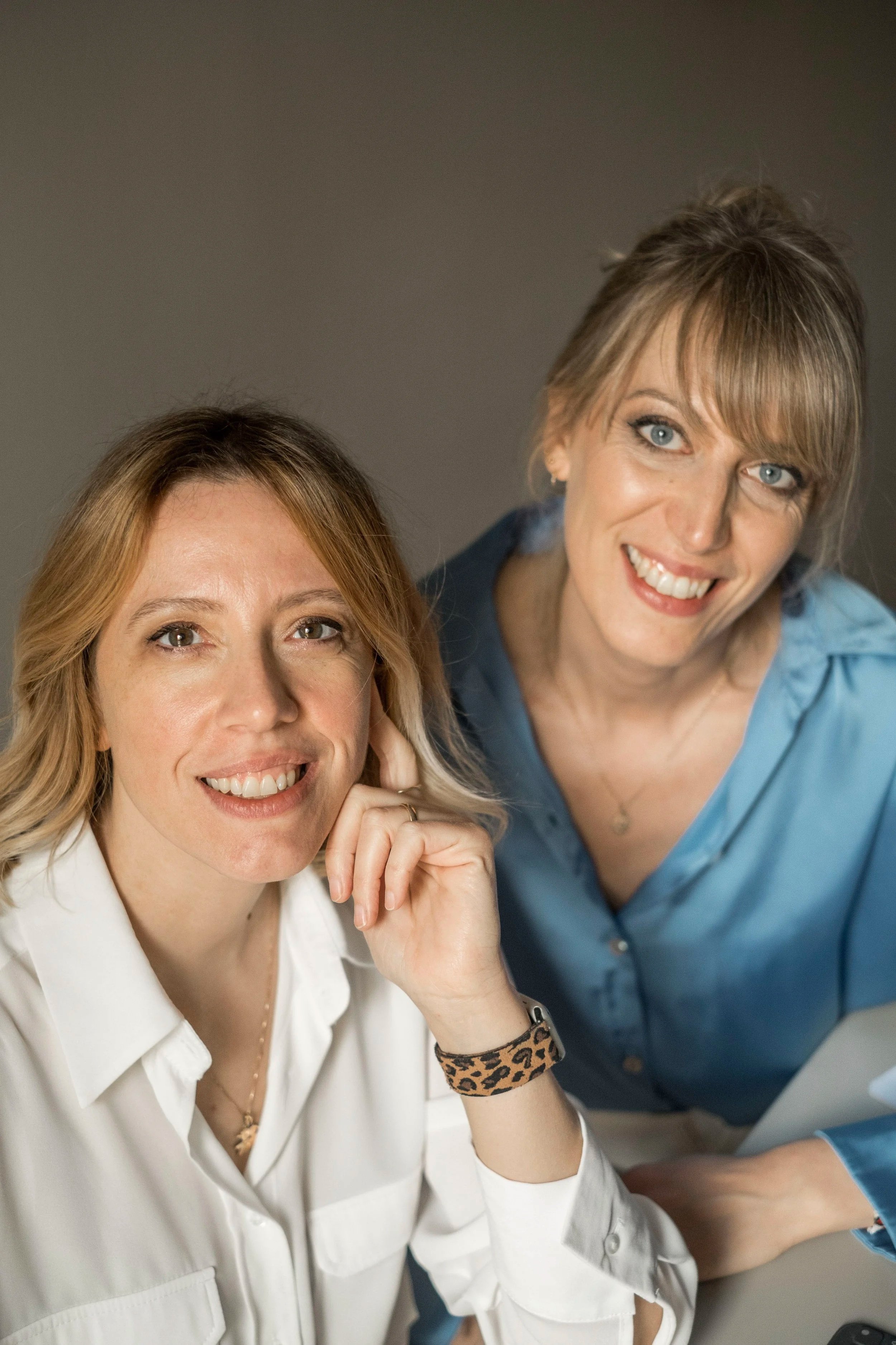 Due donne sorridenti, una con camicia bianca e l'altra con camicia azzurra, sedute vicino tra loro, in un ambiente interno, scattate in un contesto professionale o amichevole.