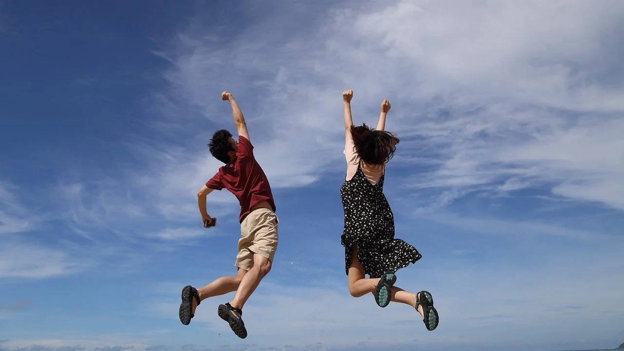 Due bambini che saltano e si sorridono sotto un cielo azzurro con nuvole sparse.