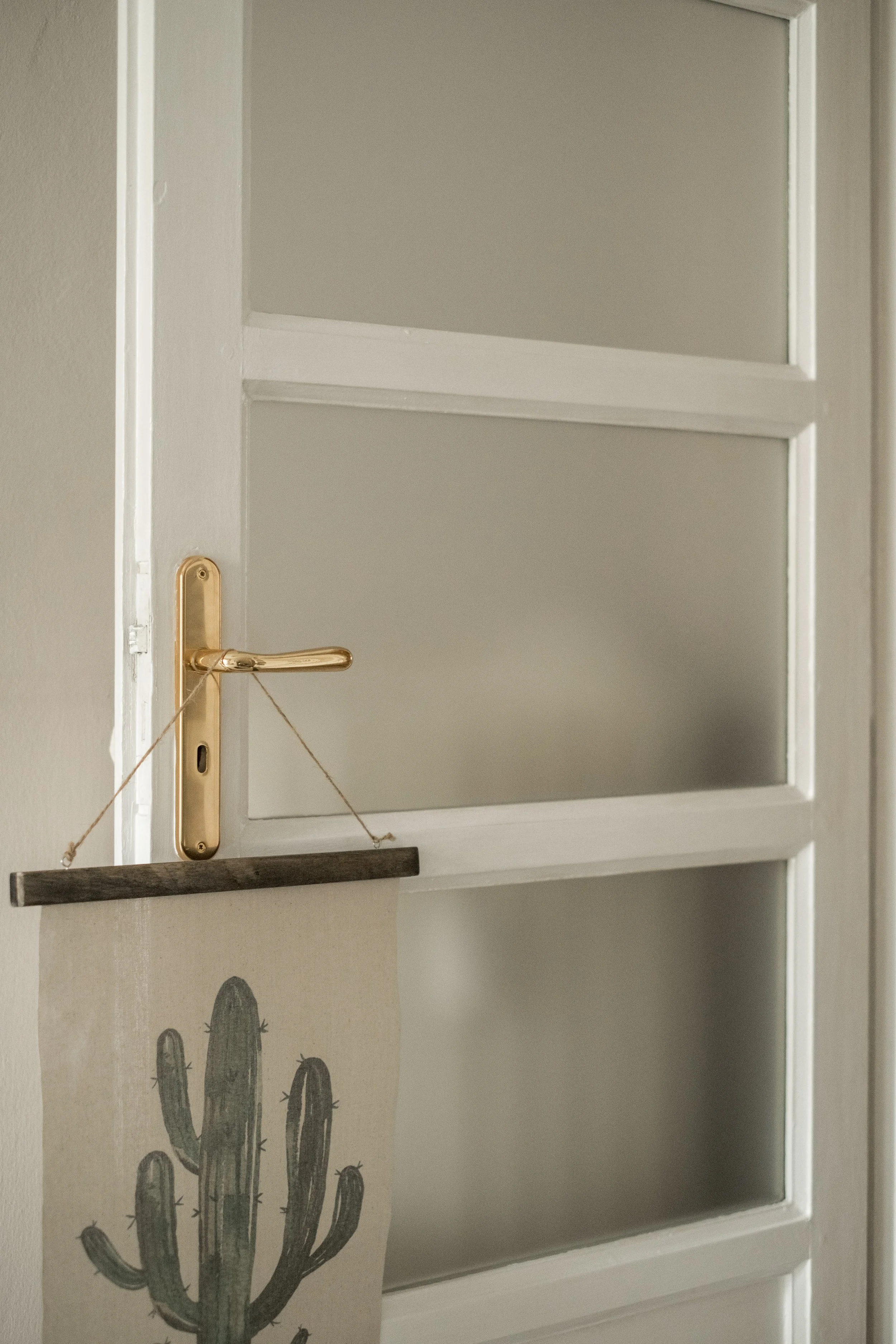 Una porta bianca con vetri traslucidi e una maniglia dorata. Sotto la maniglia, un cartello con un disegno di un cactus stilizzato appeso con una corda.
